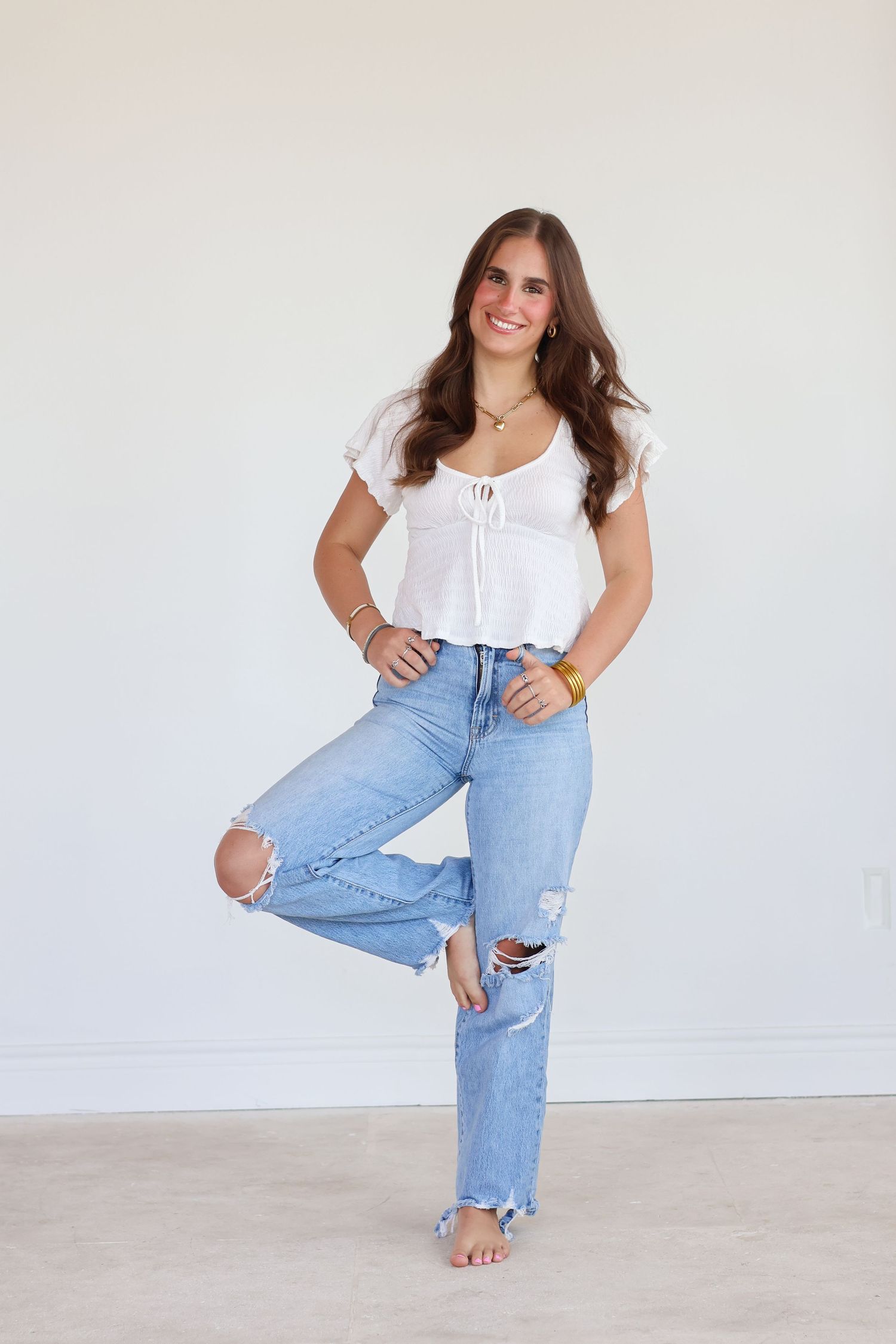 Woman in white peasant top and distressed light wash jeans poses barefoot against white wall.