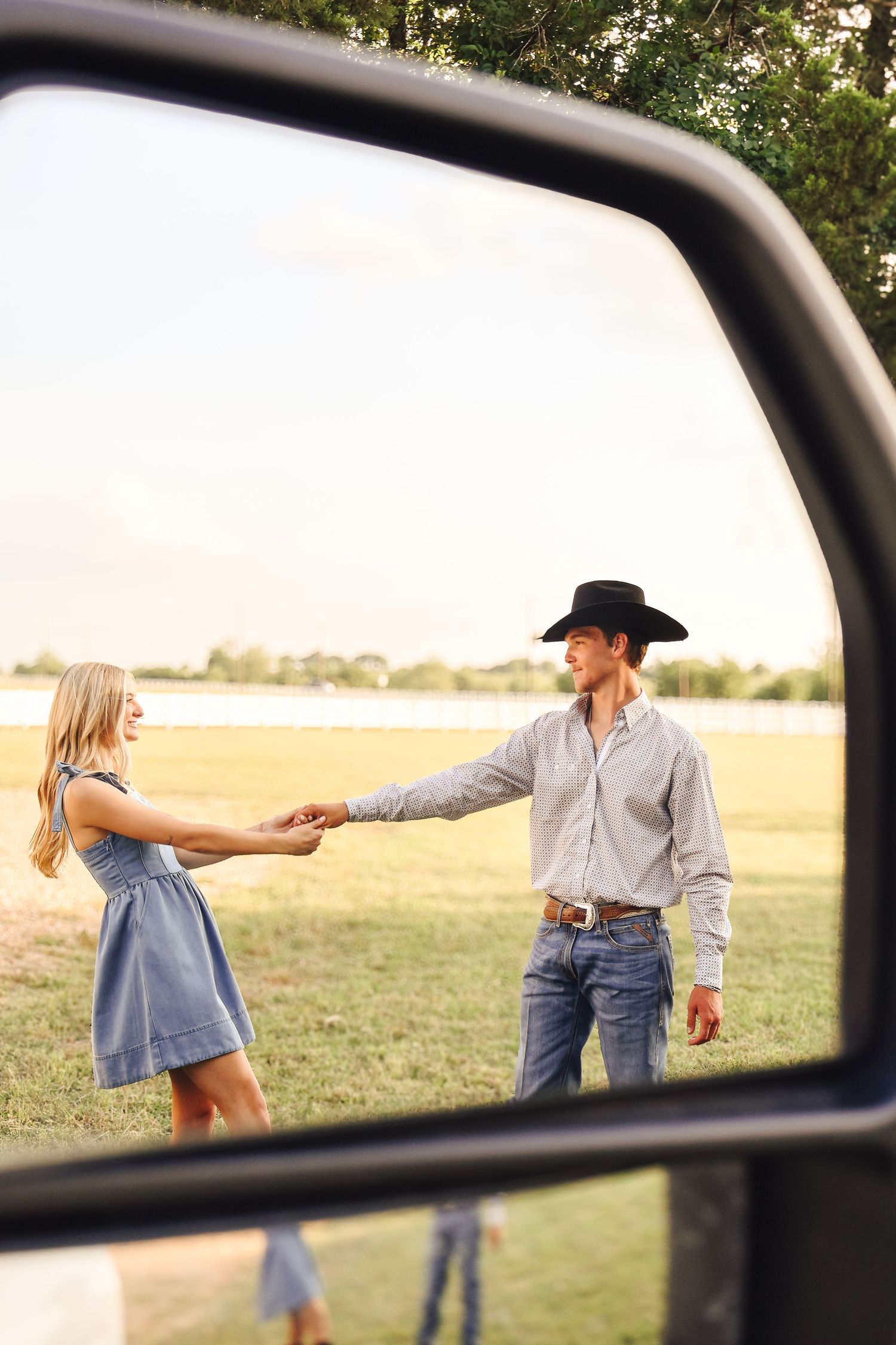 View through car window of couple dancing in an open field at sunset.