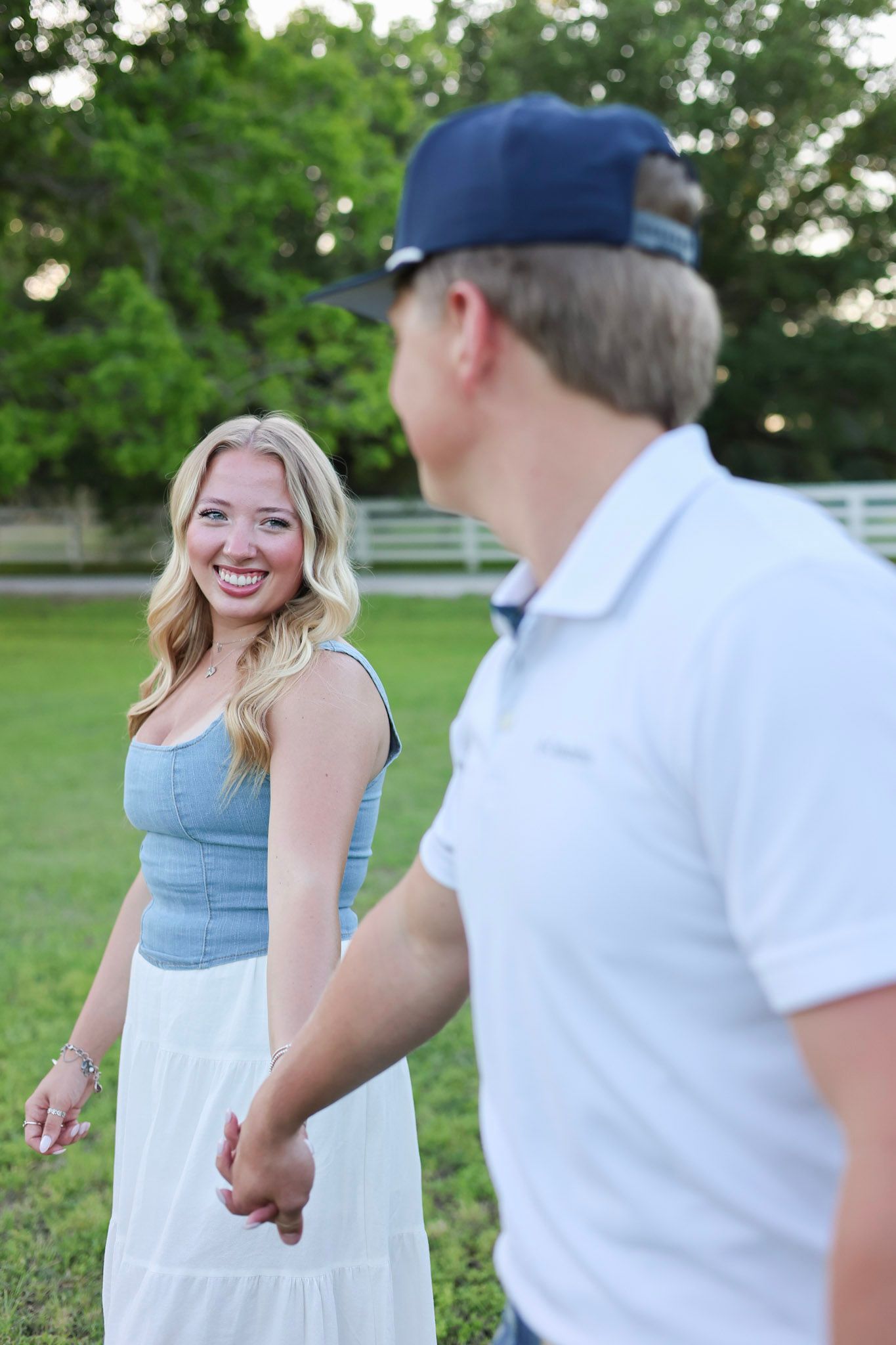 Smiling blonde woman in blue top and white skirt holds hands with man in navy cap outdoors.
