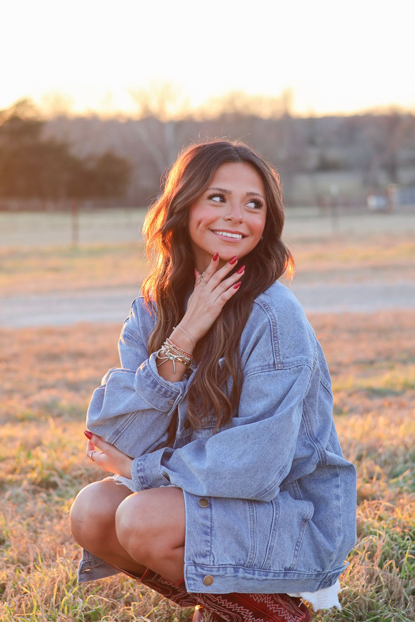 Young woman in oversized denim jacket crouching in golden field at sunset, smiling with hand on chin.