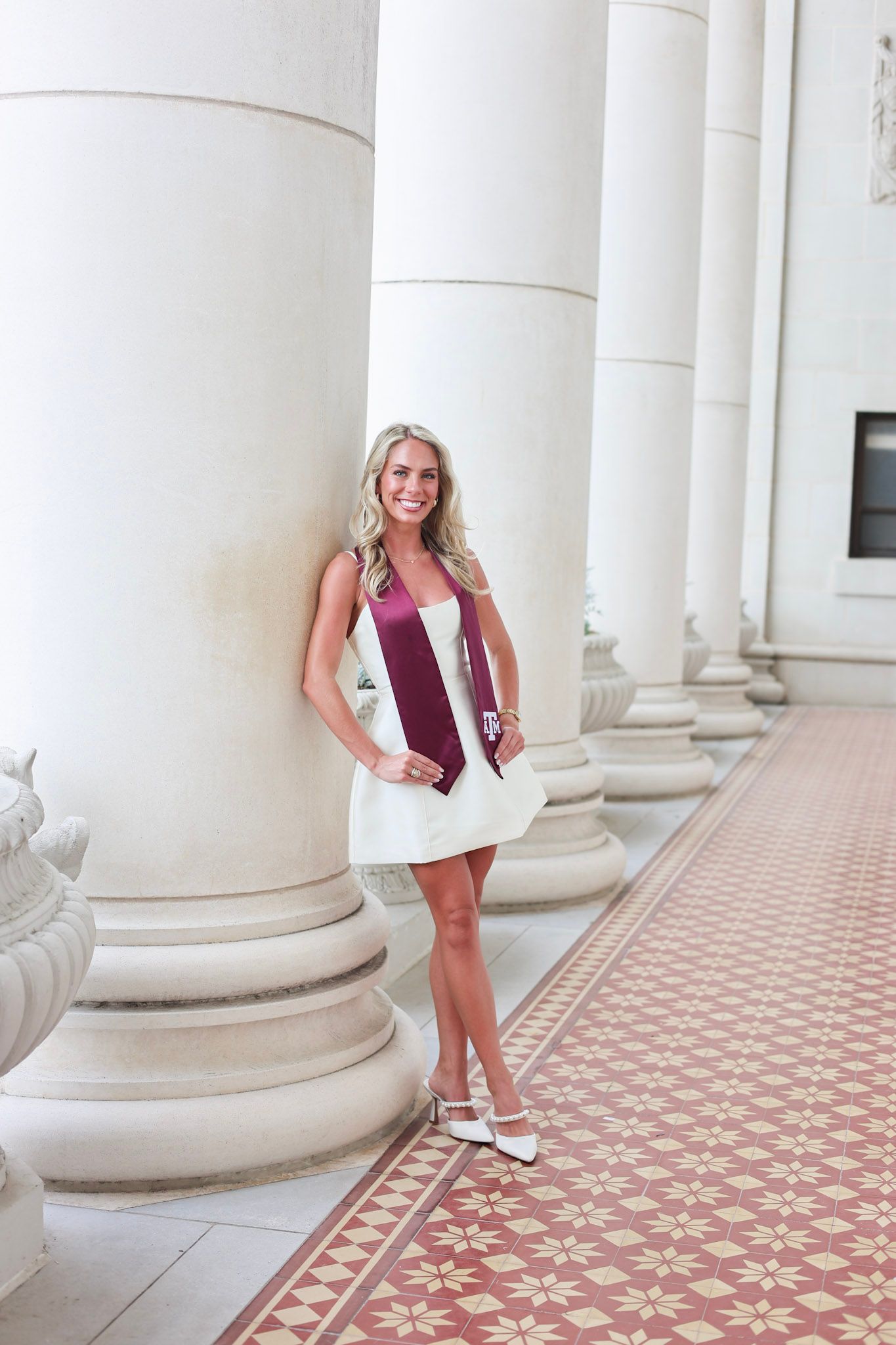 Blonde woman in white dress and Texas A&M maroon graduation stole posing between white columns on tiled floor.