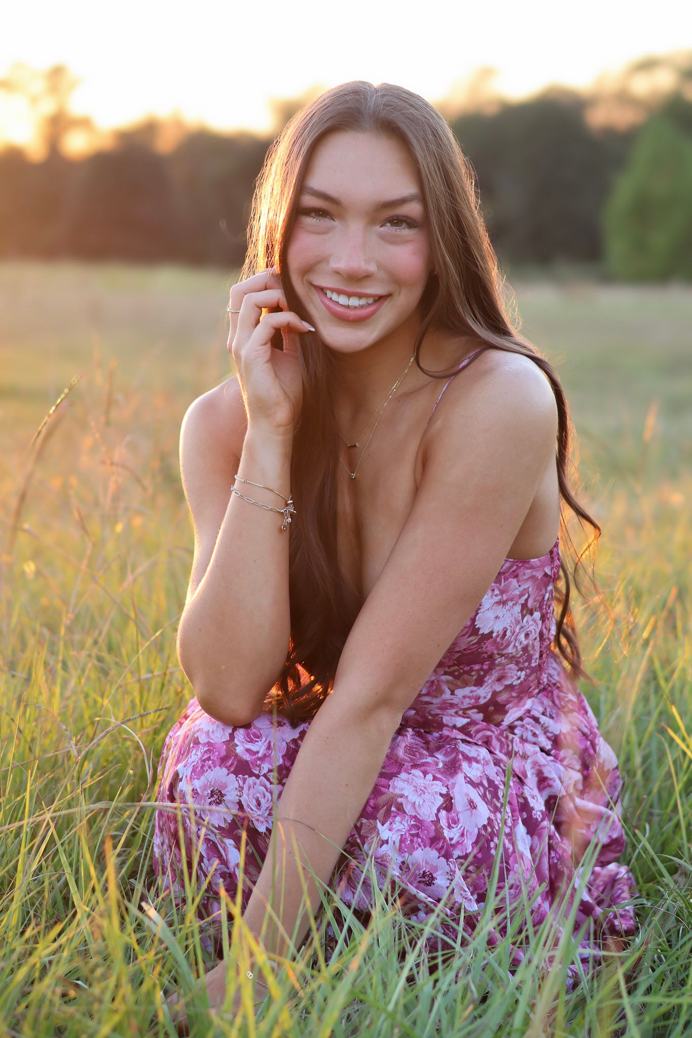 Young woman in floral dress sitting in golden hour sunlit field, smiling warmly with long brown hair.