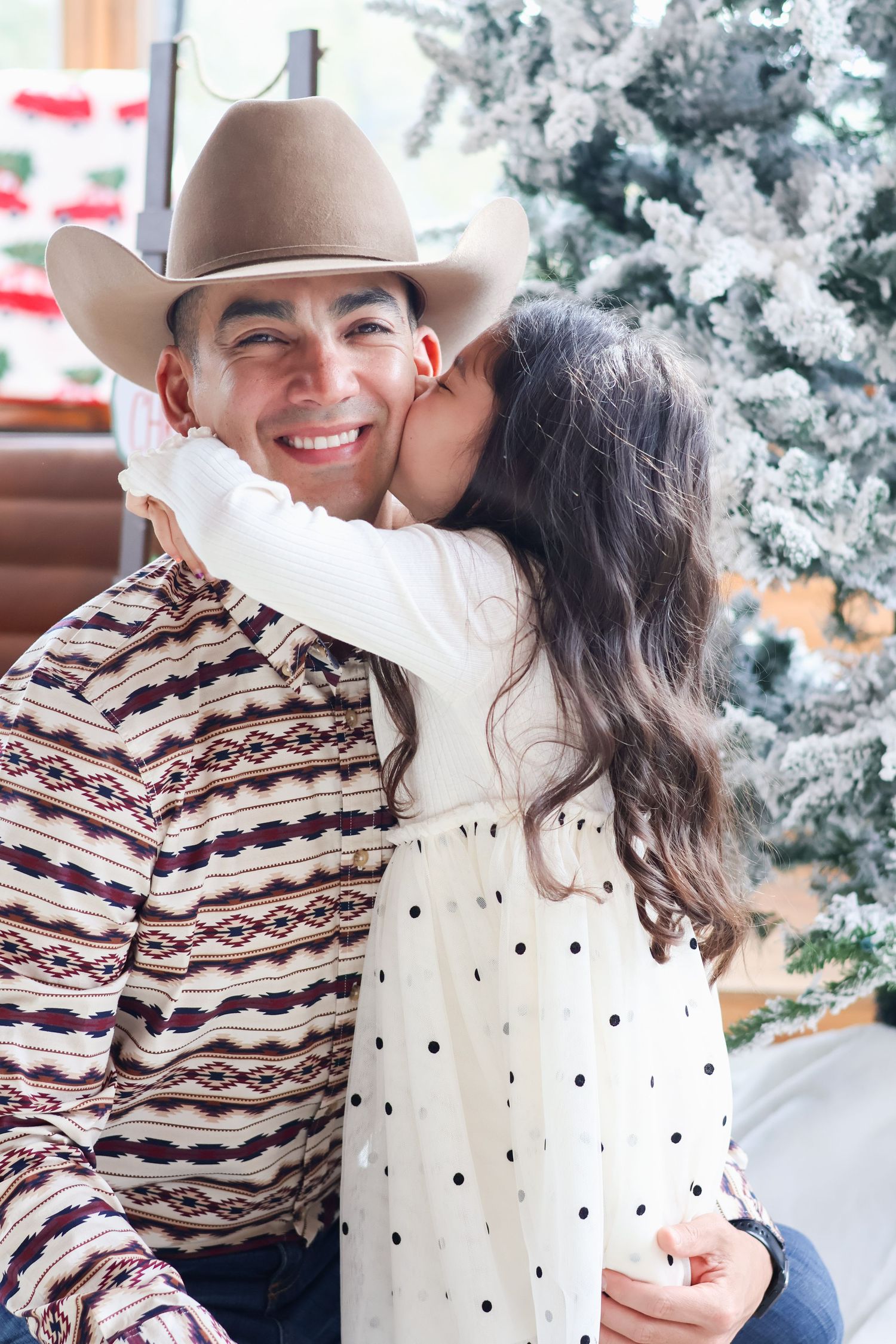 A smiling man in a cowboy hat receives a kiss on the cheek from a young girl near a Christmas tree.