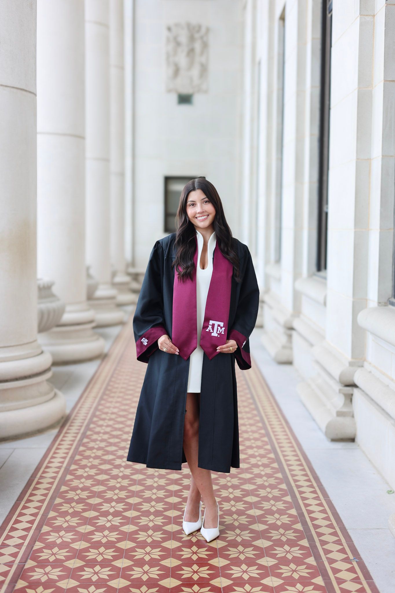 Woman in Texas A&M graduation gown and maroon stole posing in white dress and heels in columned hall.