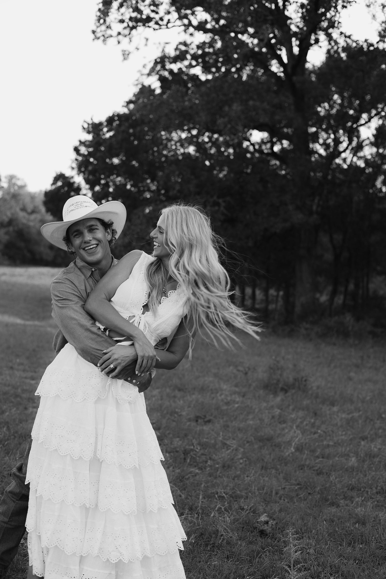 Black and white photo of couple embracing in field, man in cowboy hat, woman in white dress.