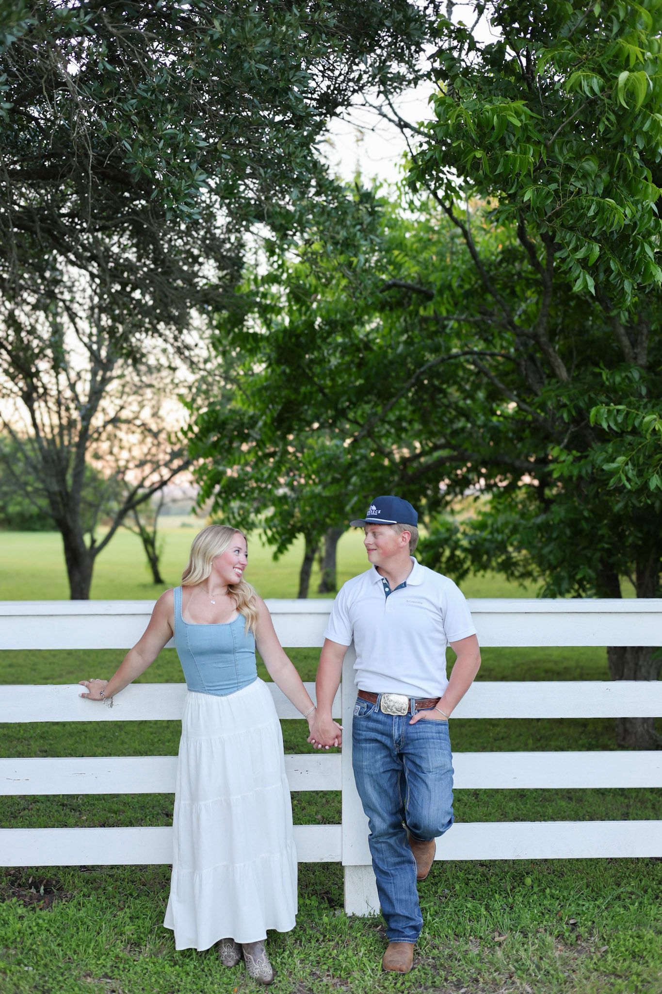 Couple holding hands by white fence, woman in blue top and white skirt, man in jeans and navy cap.