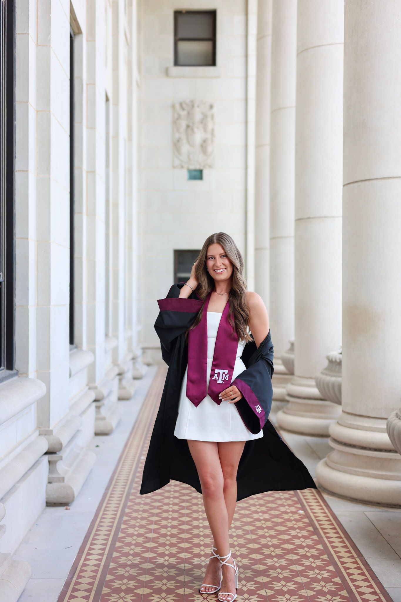 Texas A&M graduate in black gown and maroon stole poses between white columns in white dress and heels.