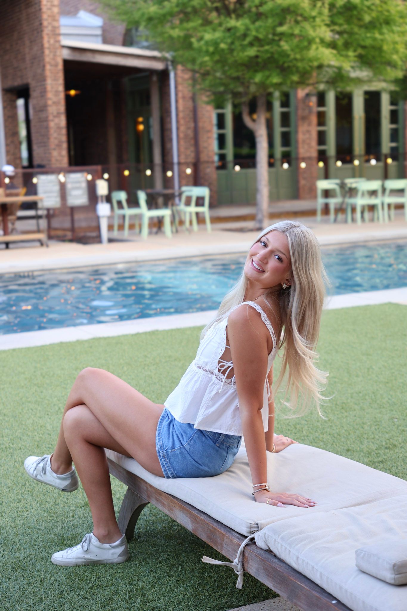Blonde woman in white top and denim shorts sitting poolside on lounge chair at outdoor venue.
