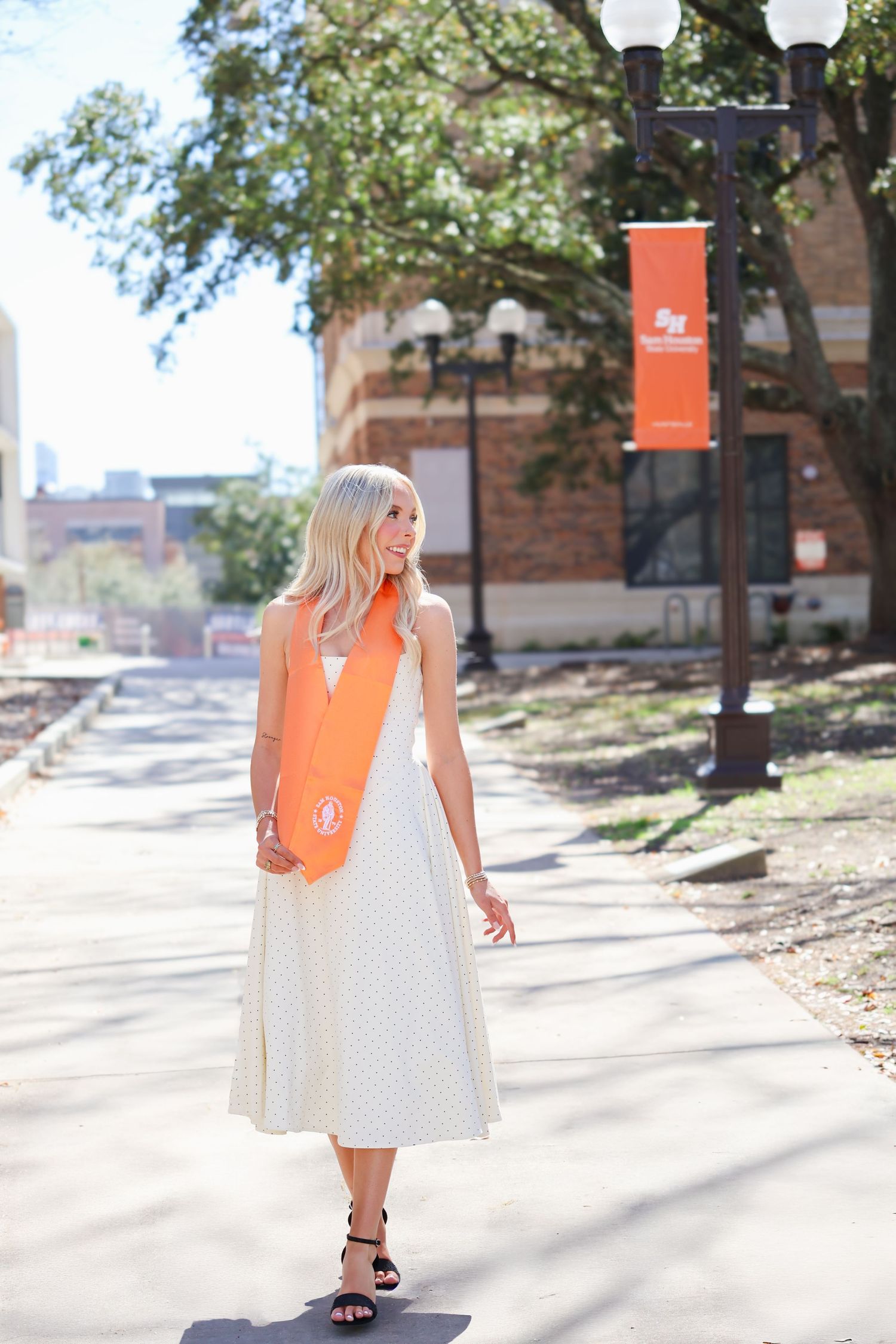 Smiling blonde woman in white dress wearing orange graduation stole walks on sunny campus pathway.