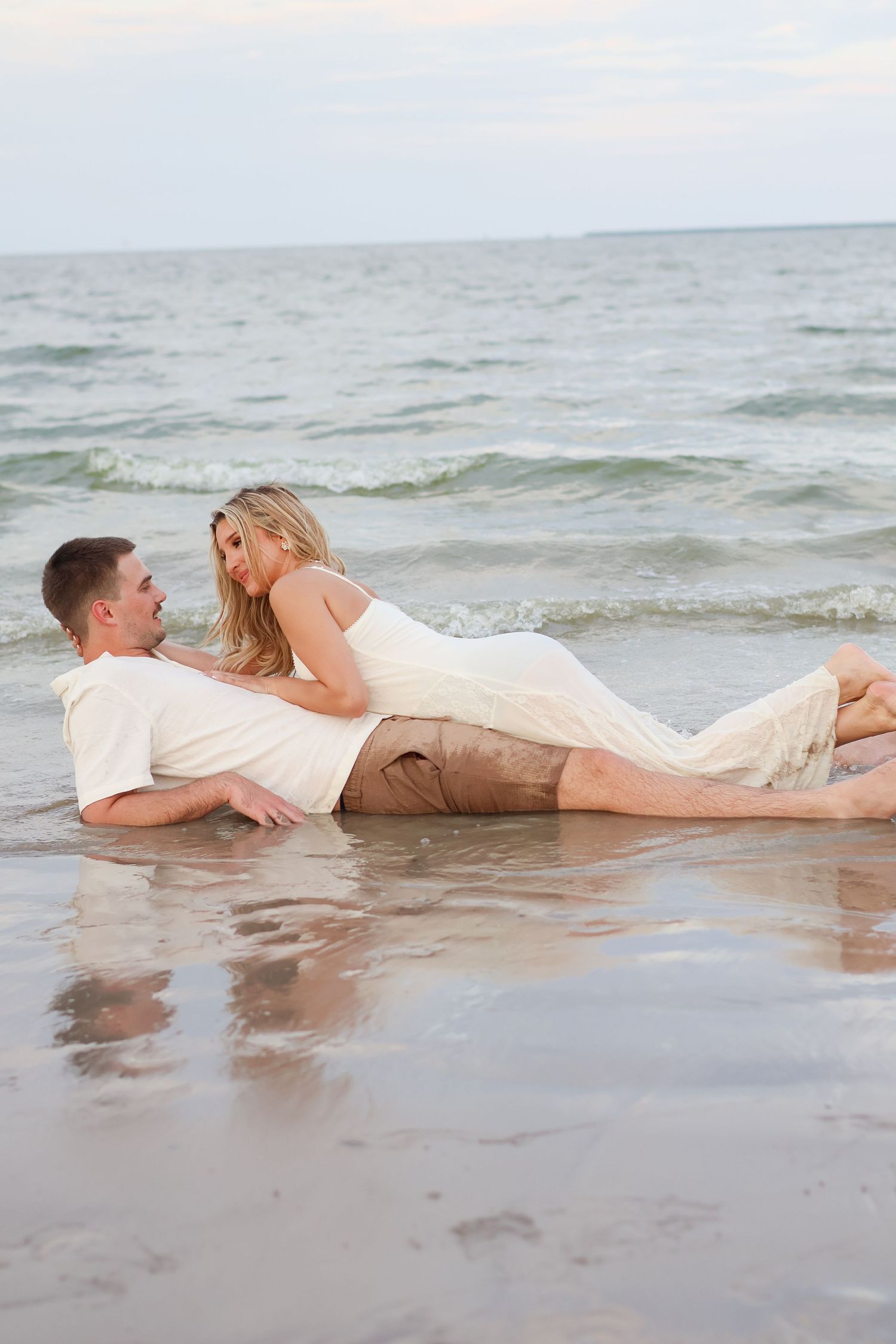 A romantic couple lies in the shallow surf at the beach wearing white and beige clothing.