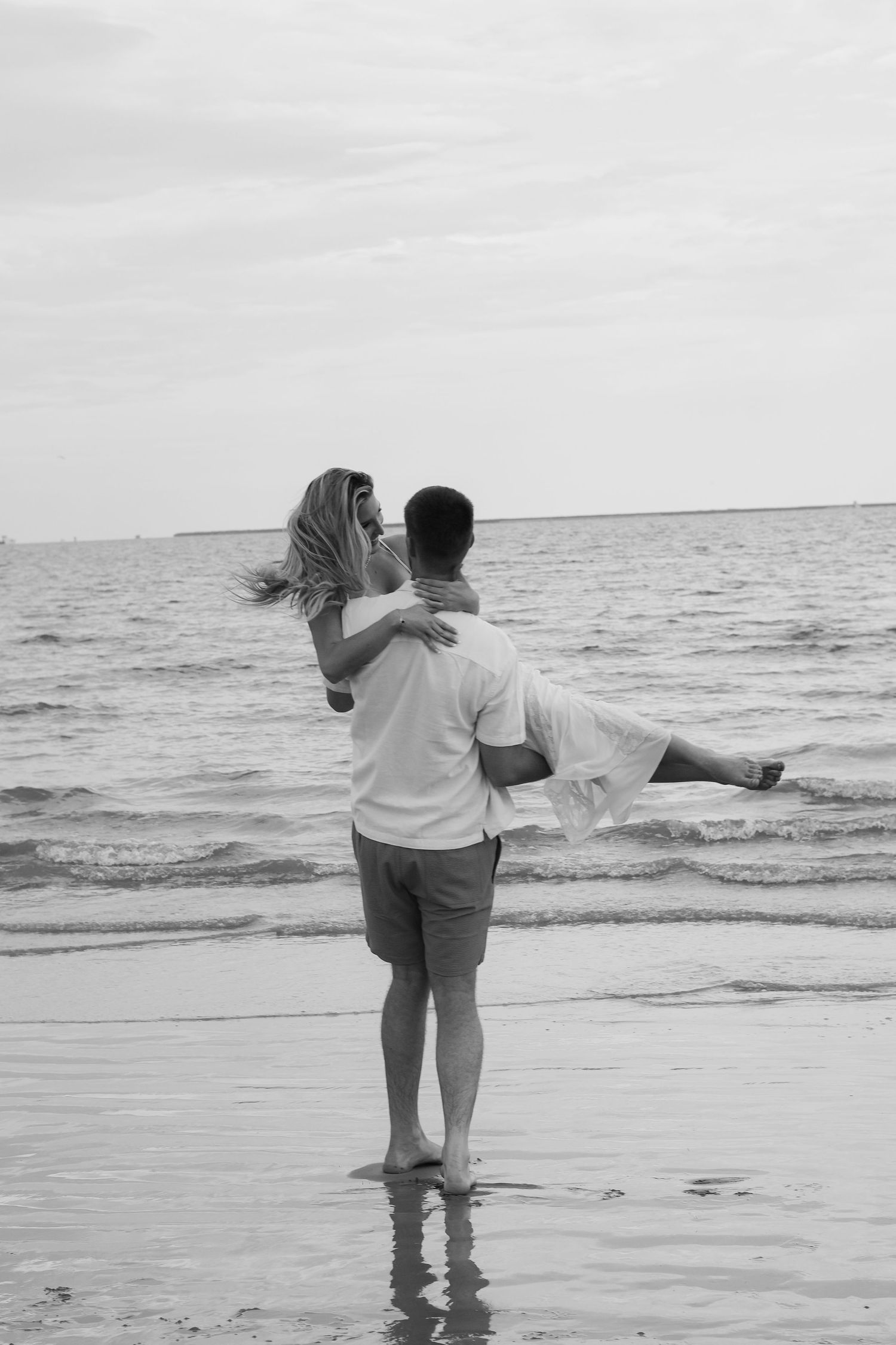 Two people stand together at the edge of the ocean waves in a romantic black and white beach scene.