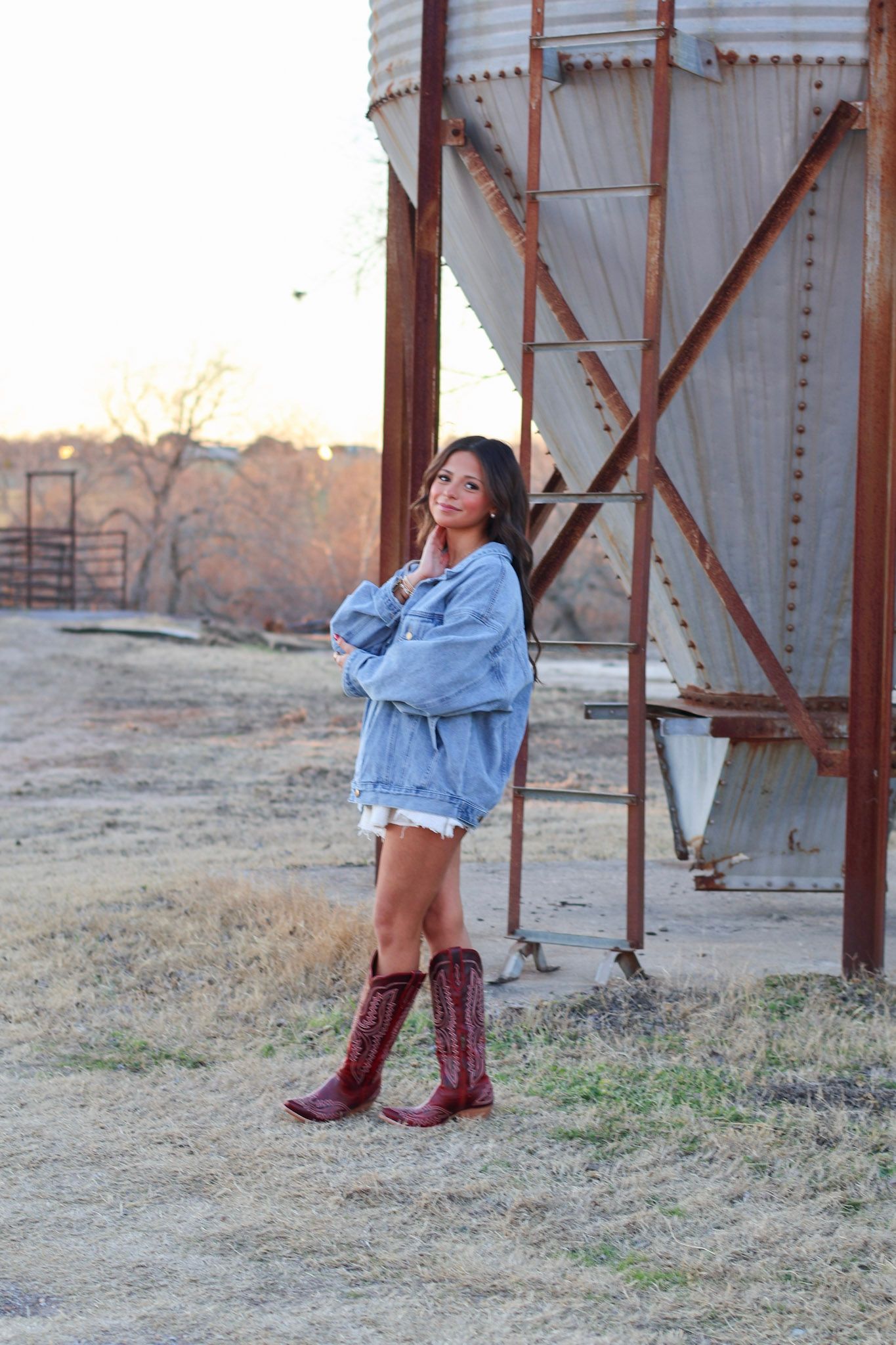 Young woman in oversized denim jacket and red cowboy boots posing near rustic grain silo at sunset.