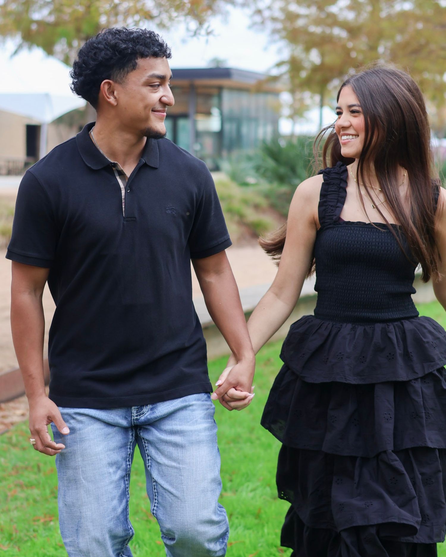 A young couple holding hands and smiling while walking on green grass, both wearing matching black outfits.