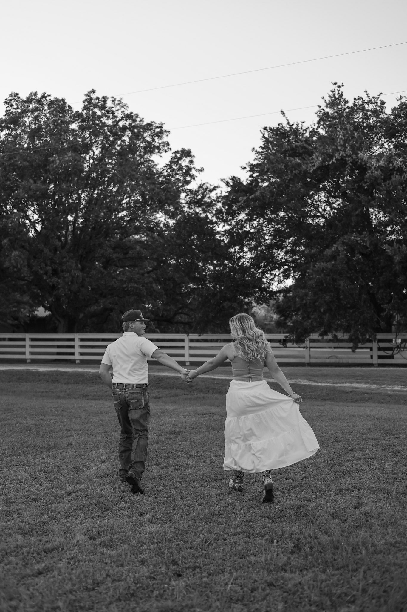 Black and white photo of a bride and groom holding hands walking through a grassy field toward a white fence.
