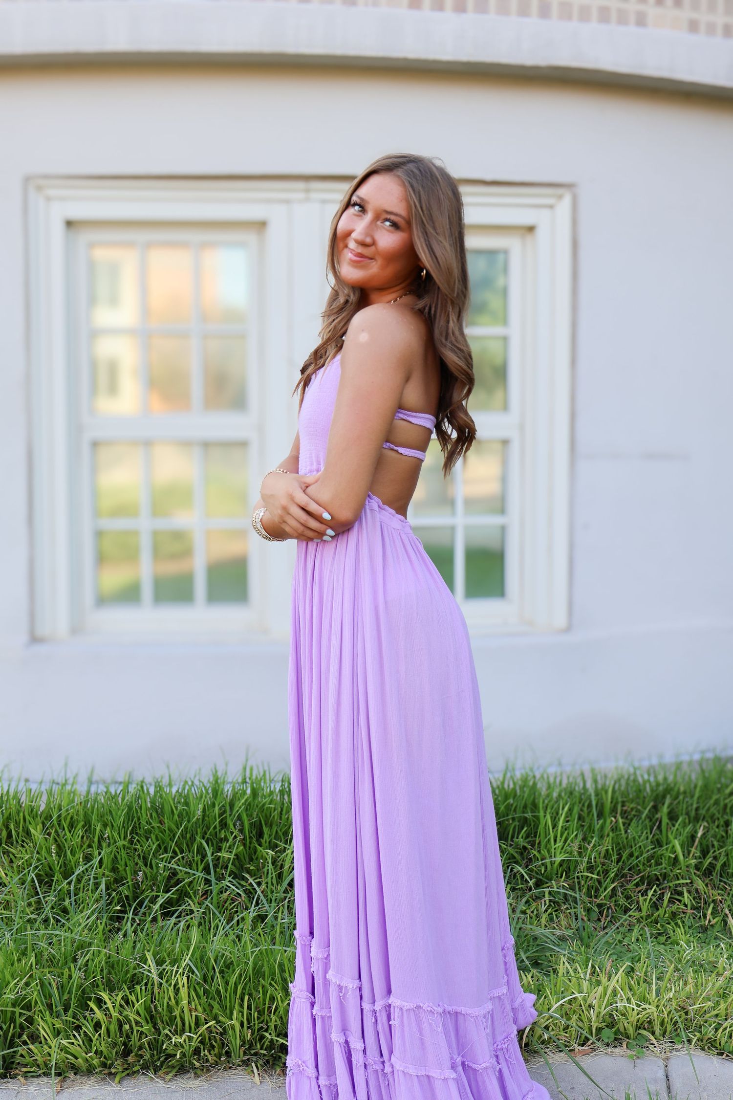 Woman in flowing purple maxi dress poses beside white building.