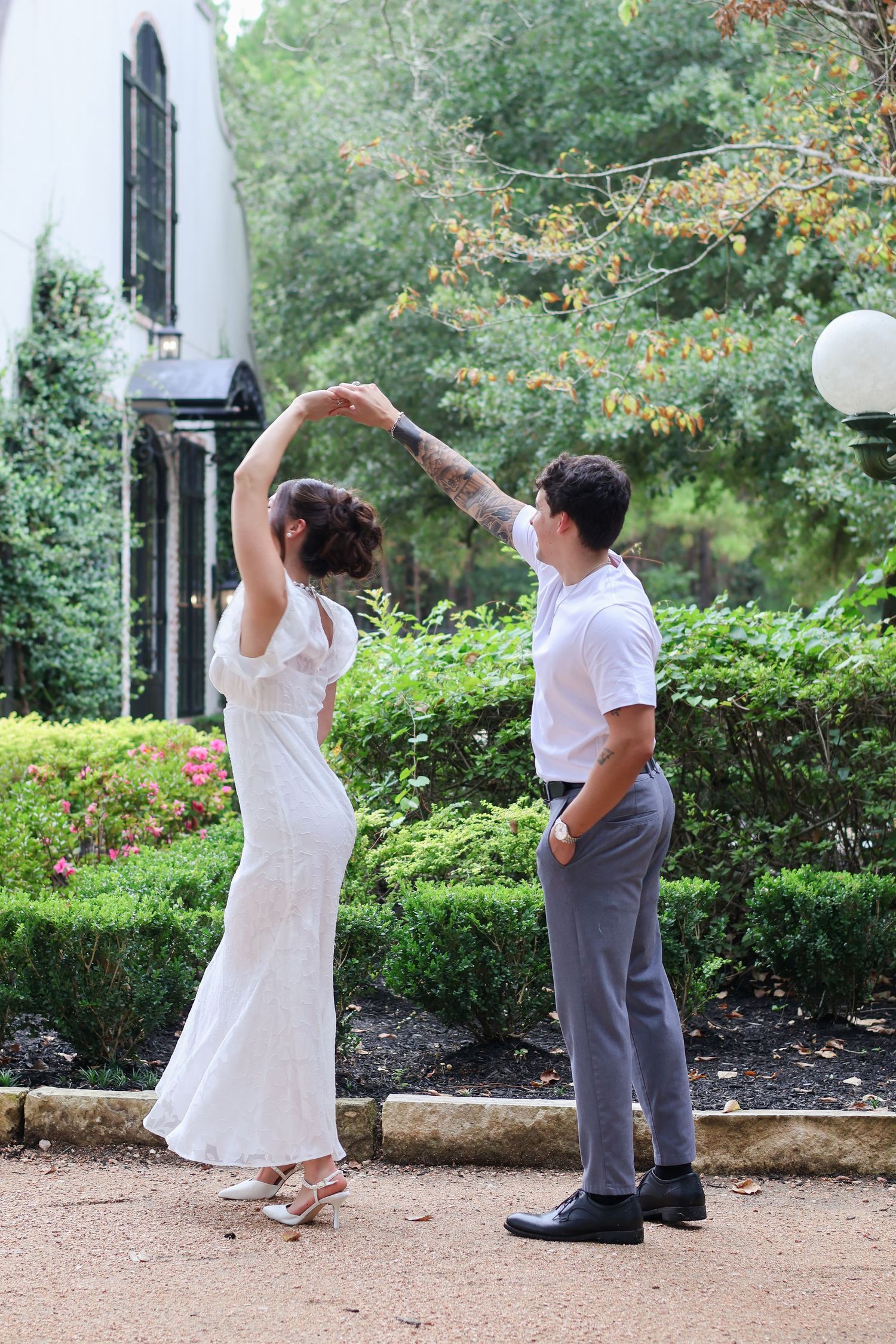 Two people in white and gray outfits perform a fun dance sequence in a garden setting with greenery in the background.