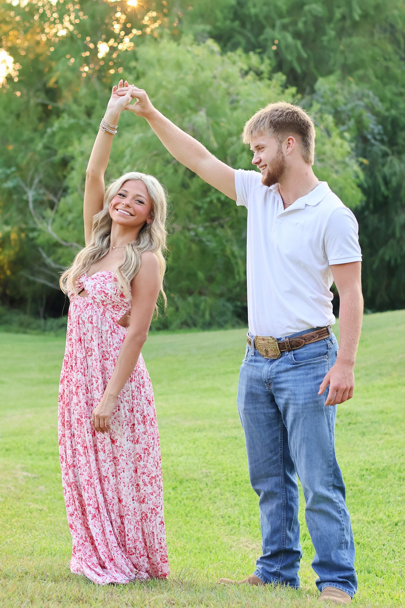 A couple dances together on a grassy lawn at sunset, with her in a pink floral maxi dress and him in jeans and white shirt.