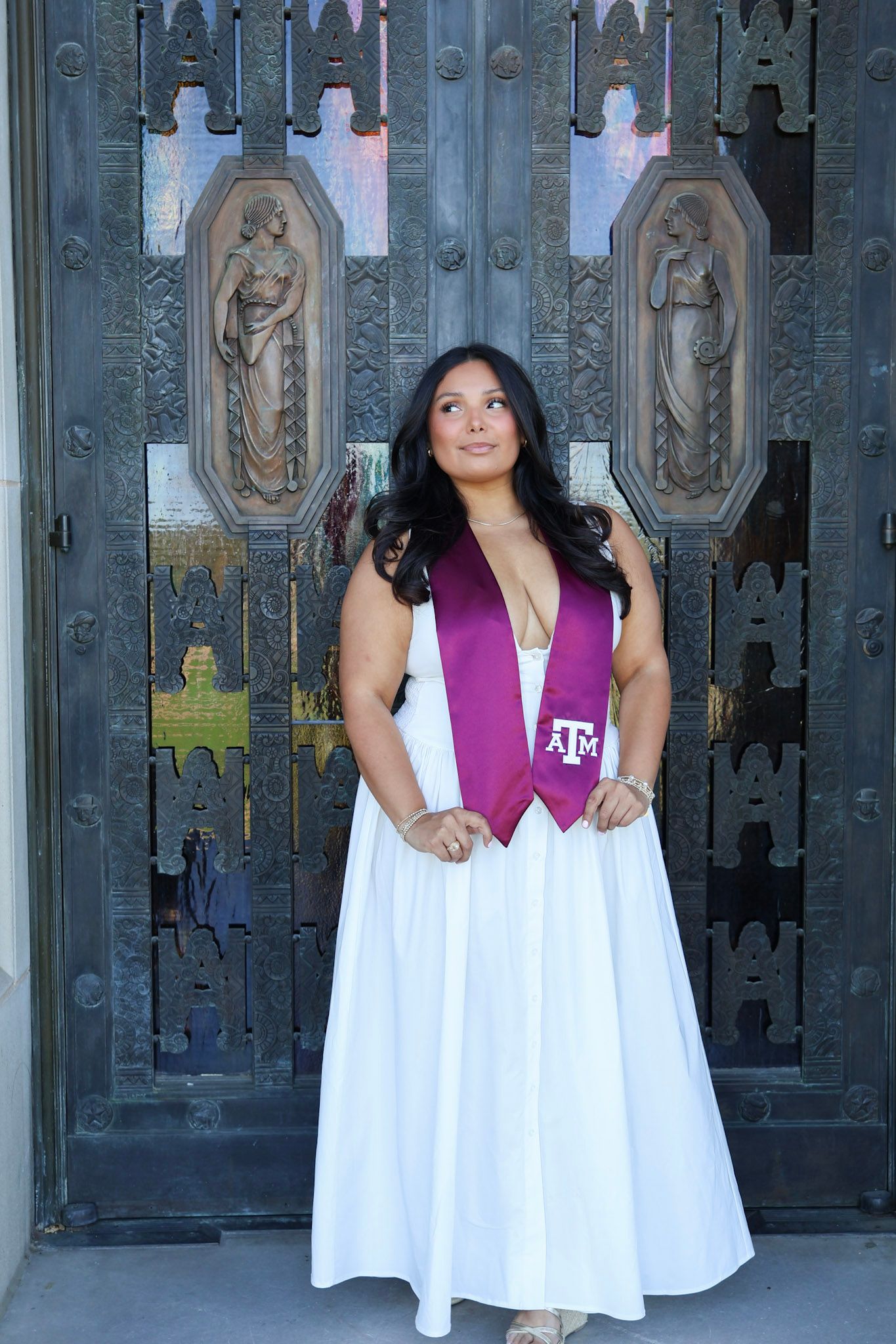 Woman in white gown with Texas A&M maroon graduation stole poses before ornate bronze doors.