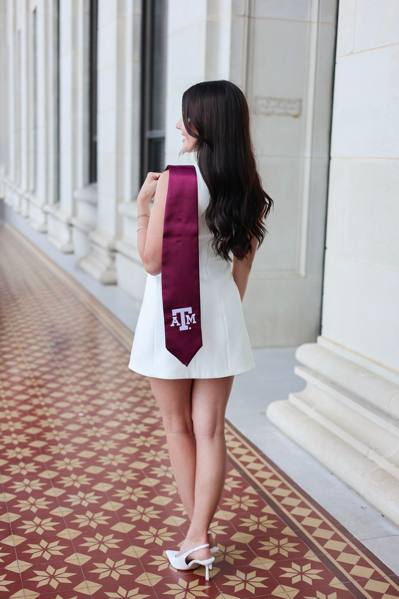 Young woman in white dress wearing Texas A&M maroon graduation stole, posing in elegant columned hallway.