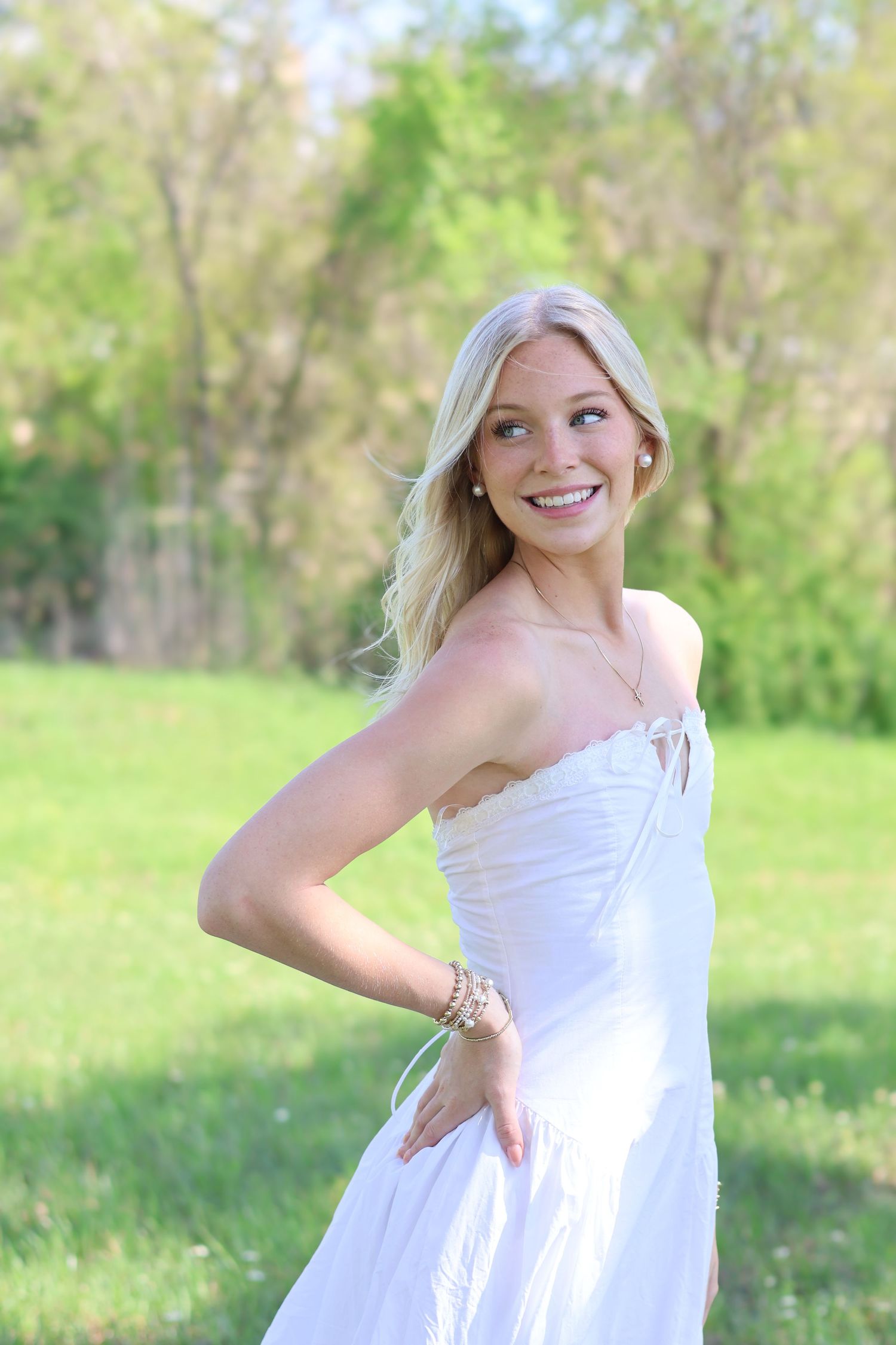 Smiling blonde woman in white strapless dress posing outdoors in green park setting.