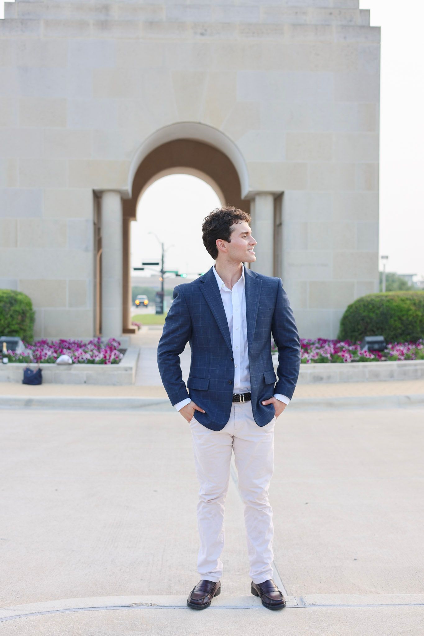 Young man in navy blazer and white pants stands confidently before a stone archway monument with floral gardens.