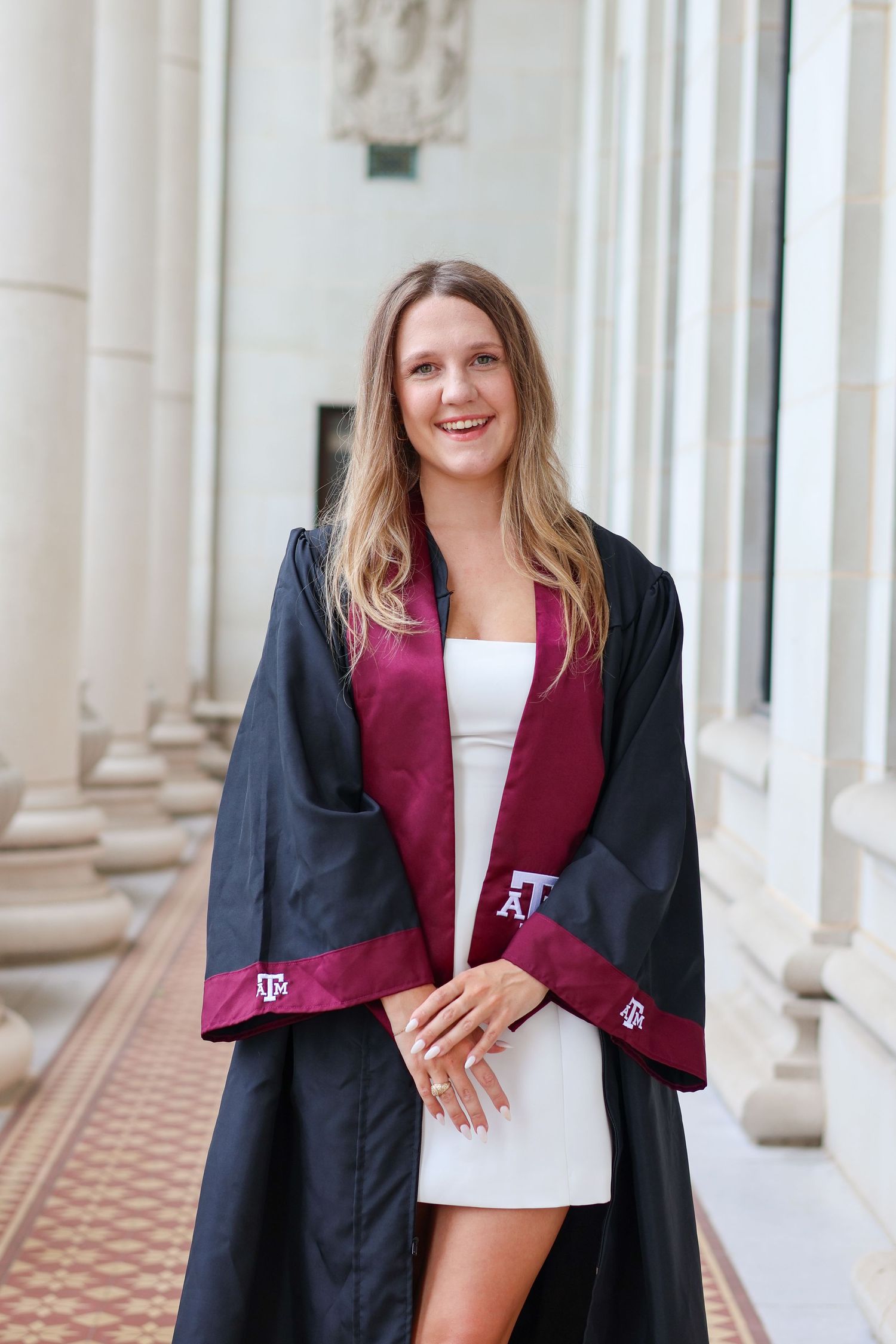Graduate in burgundy and black academic regalia poses for celebratory photos in a classical columned hallway.