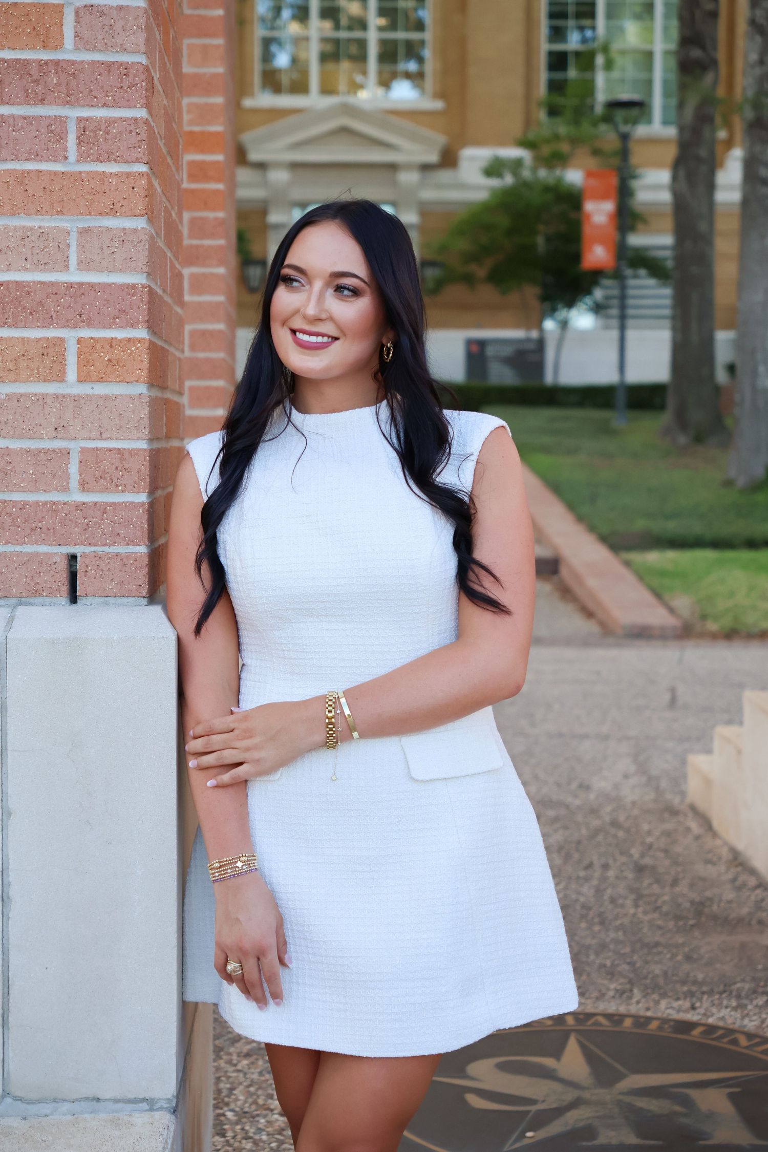 A woman in a white sleeveless top poses confidently against a brick wall background.