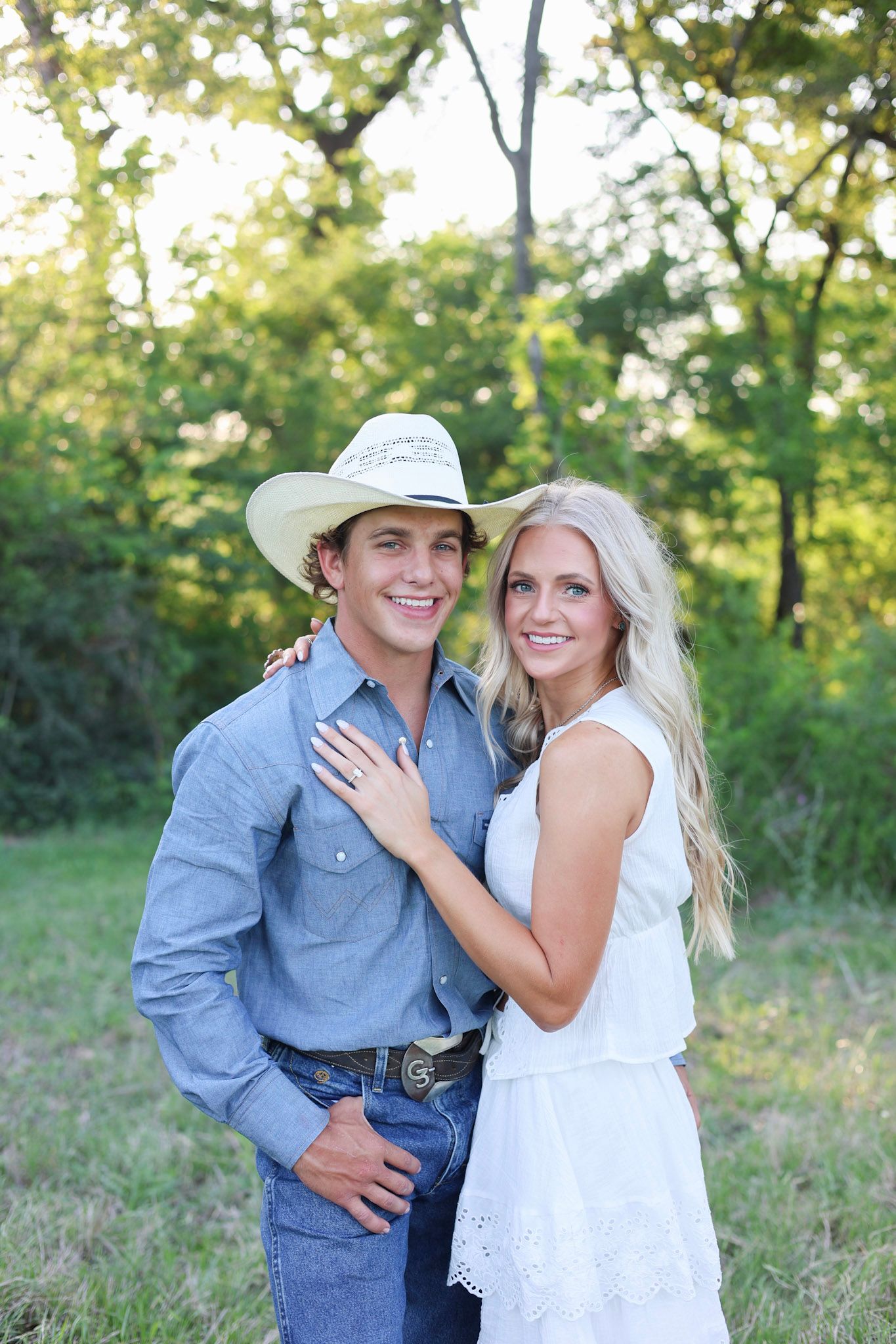 Smiling couple posing outdoors, man in cowboy hat and blue shirt, woman in white dress.