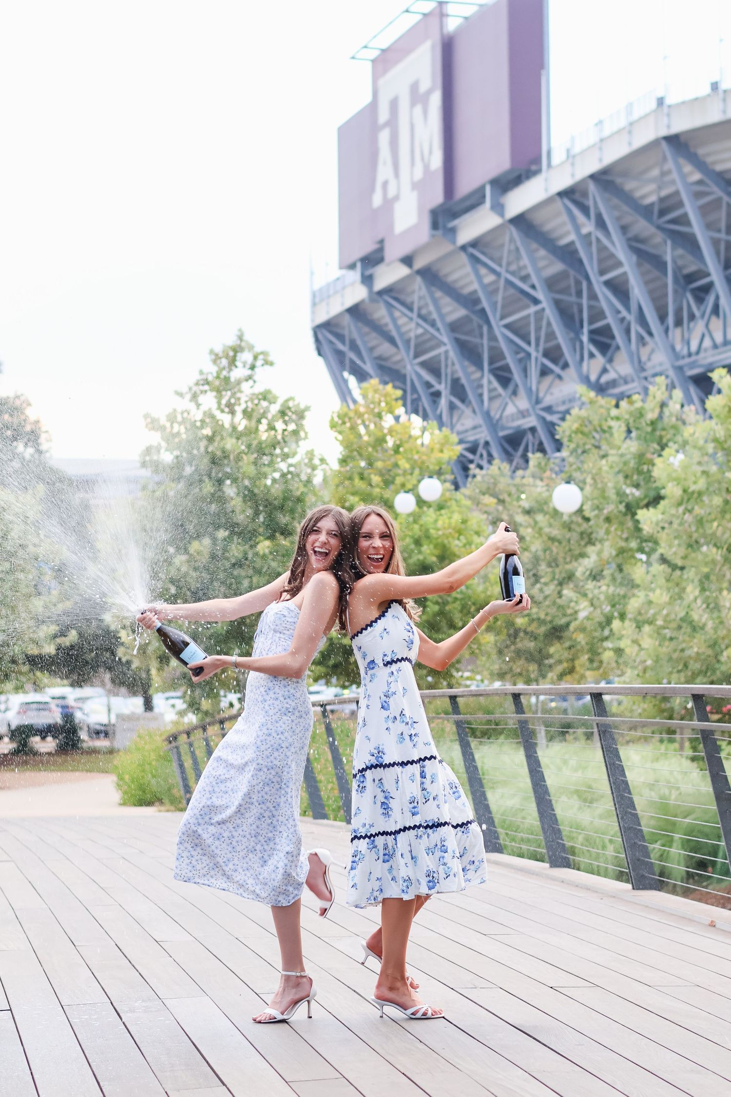 Two people in matching blue and white dresses pose playfully on a wooden deck with stadium architecture visible behind them.