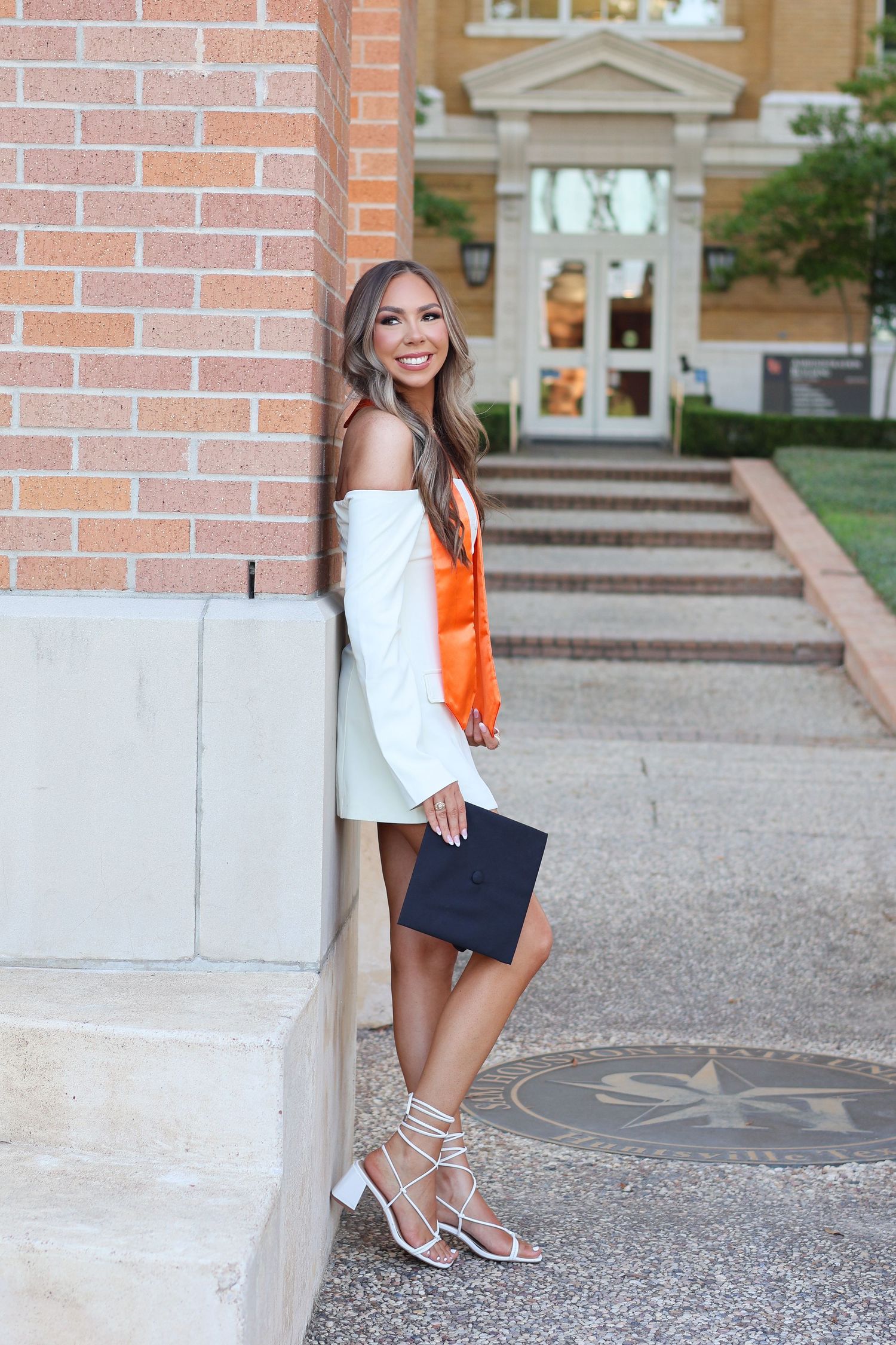 A person in an orange top and navy shorts poses on brick steps near a white column.