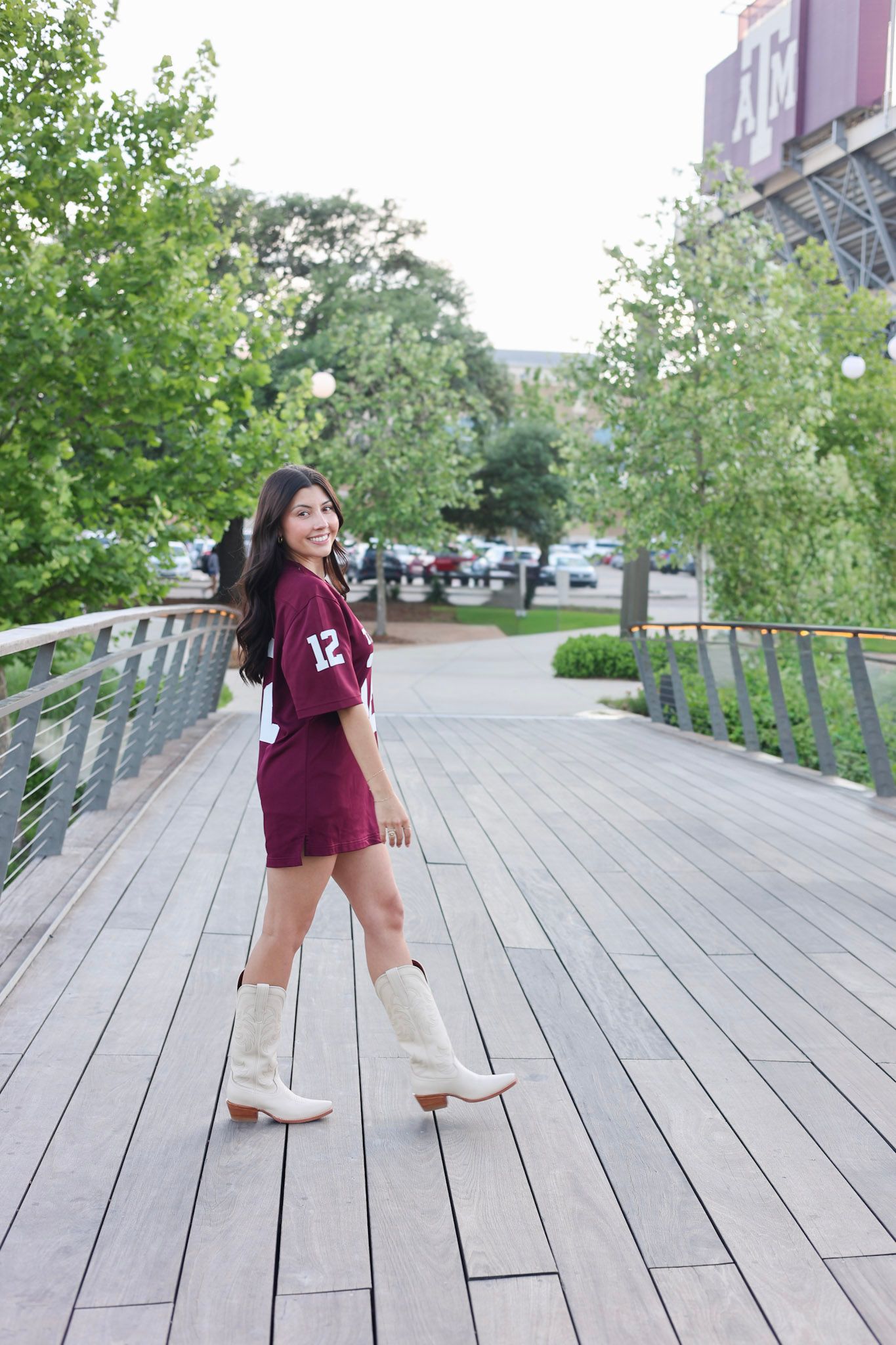 Brunette woman in maroon Texas A&M jersey dress and white cowboy boots on wooden walkway near stadium.