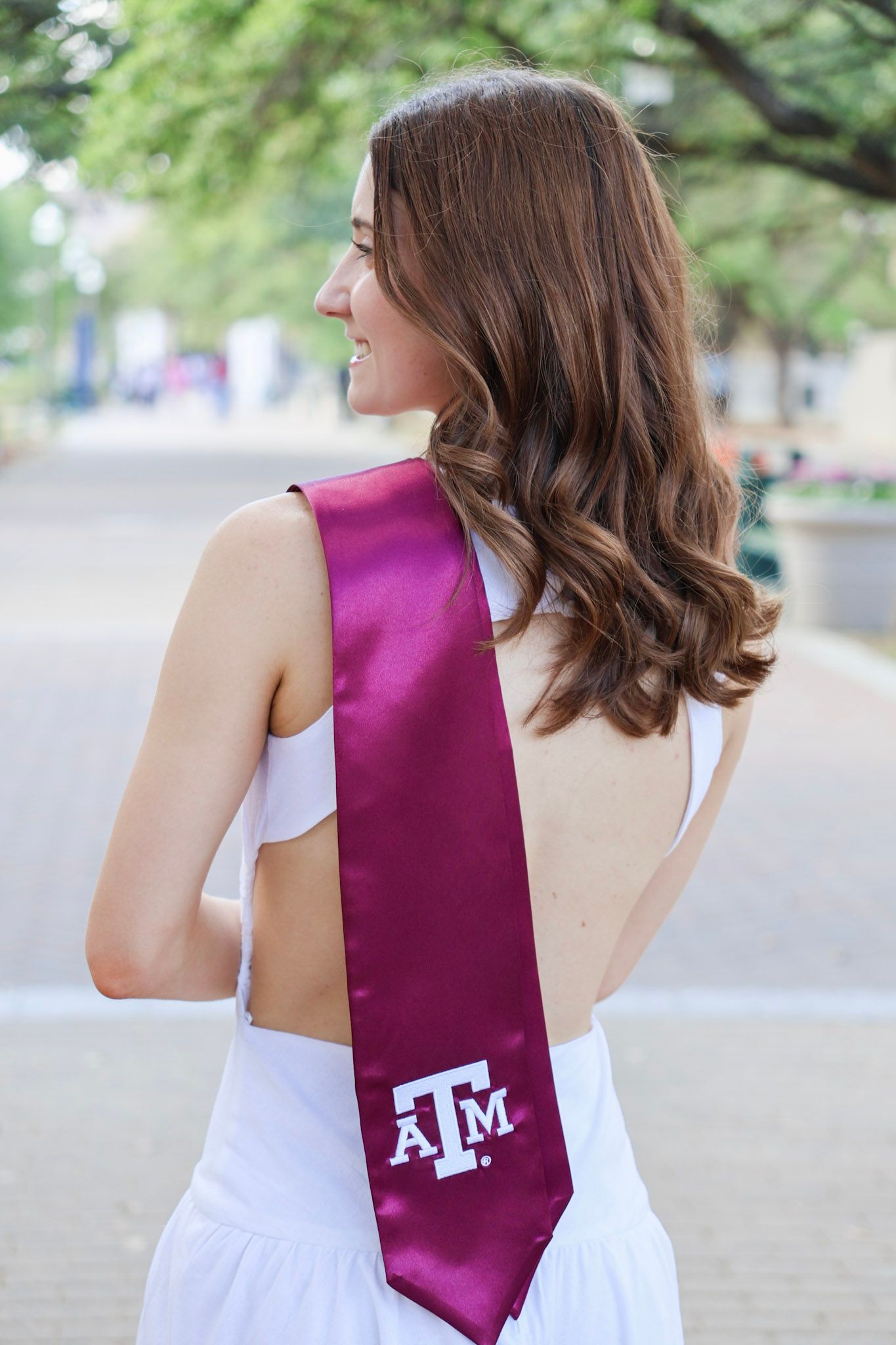 Young woman wearing a white dress and maroon Texas A&M University graduation sash, smiling outdoors on campus.