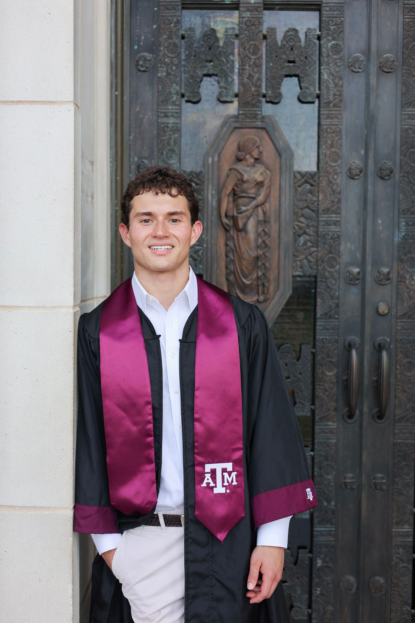College graduate in Texas A&M maroon and black graduation regalia standing by ornate bronze doors.