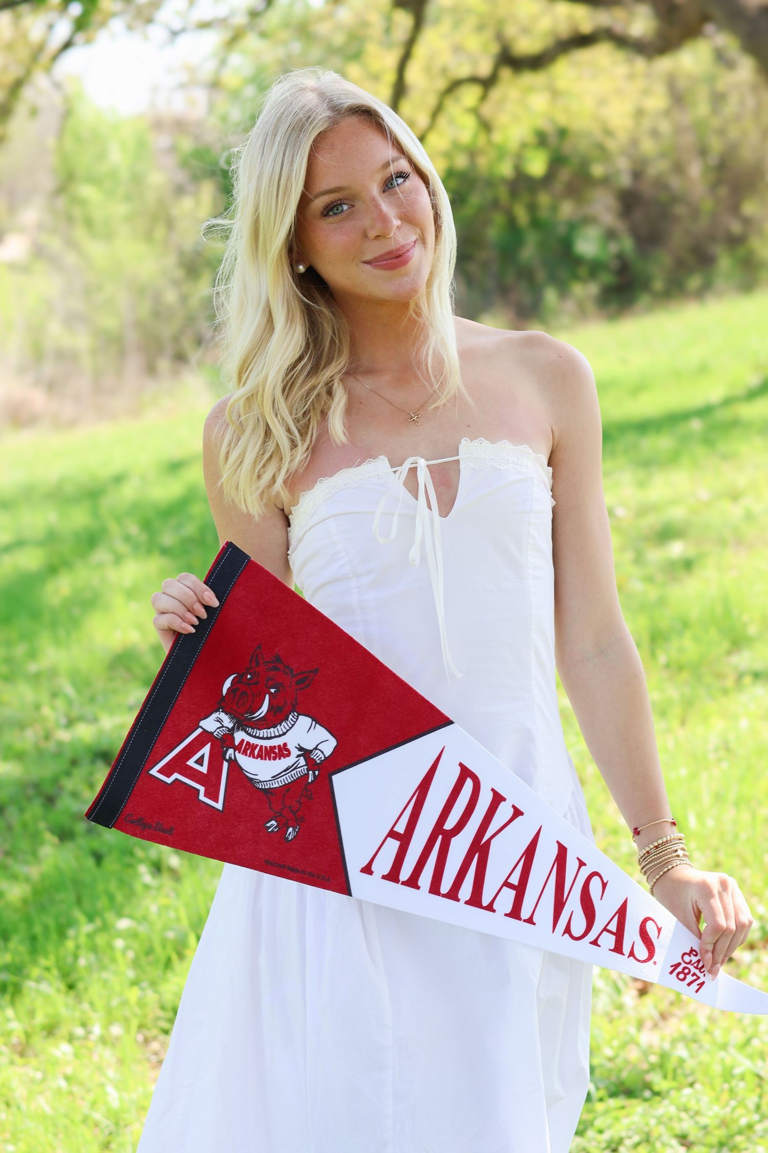 Blonde woman in white dress holding Arkansas Razorbacks pennant flag outdoors on green grass.
