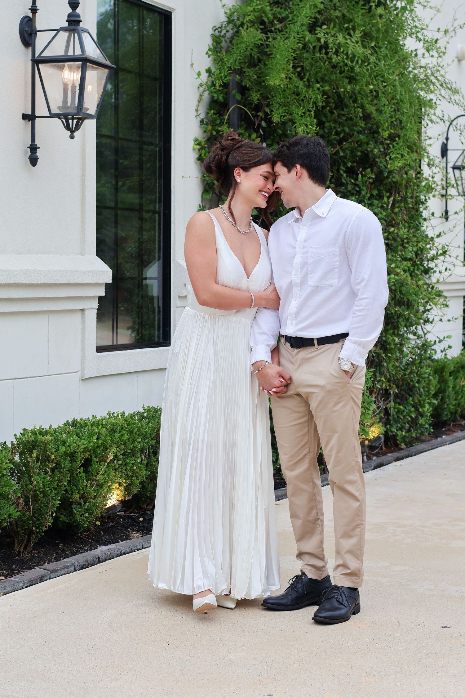 A couple in white and khaki outfits share an intimate moment against a white building with green foliage.