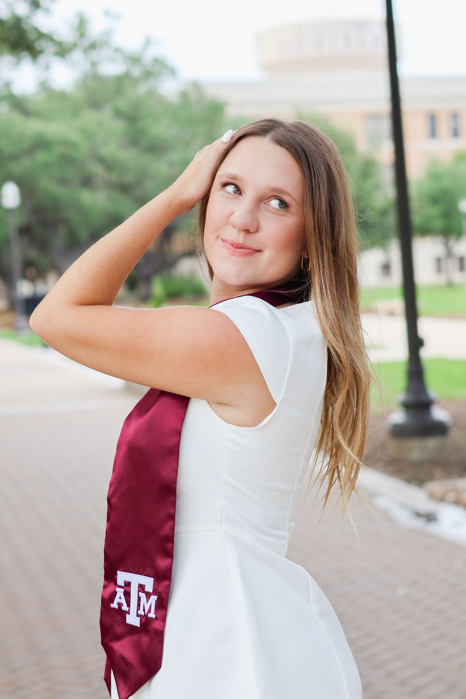 A student poses playfully in a white top and Texas A&M maroon shirt on a university campus walkway.