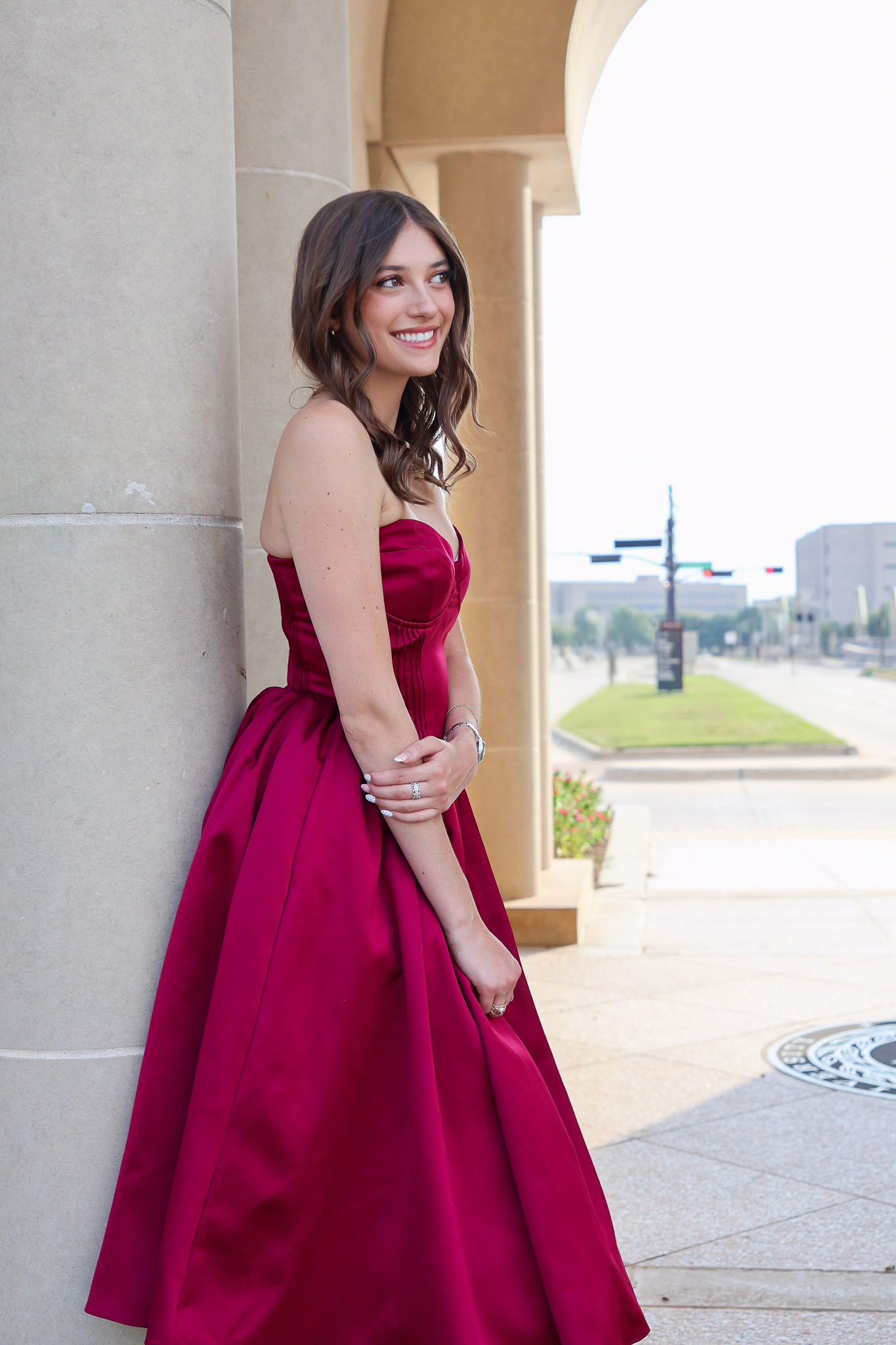 A person in a burgundy strapless ball gown poses elegantly against stone columns in an outdoor setting.