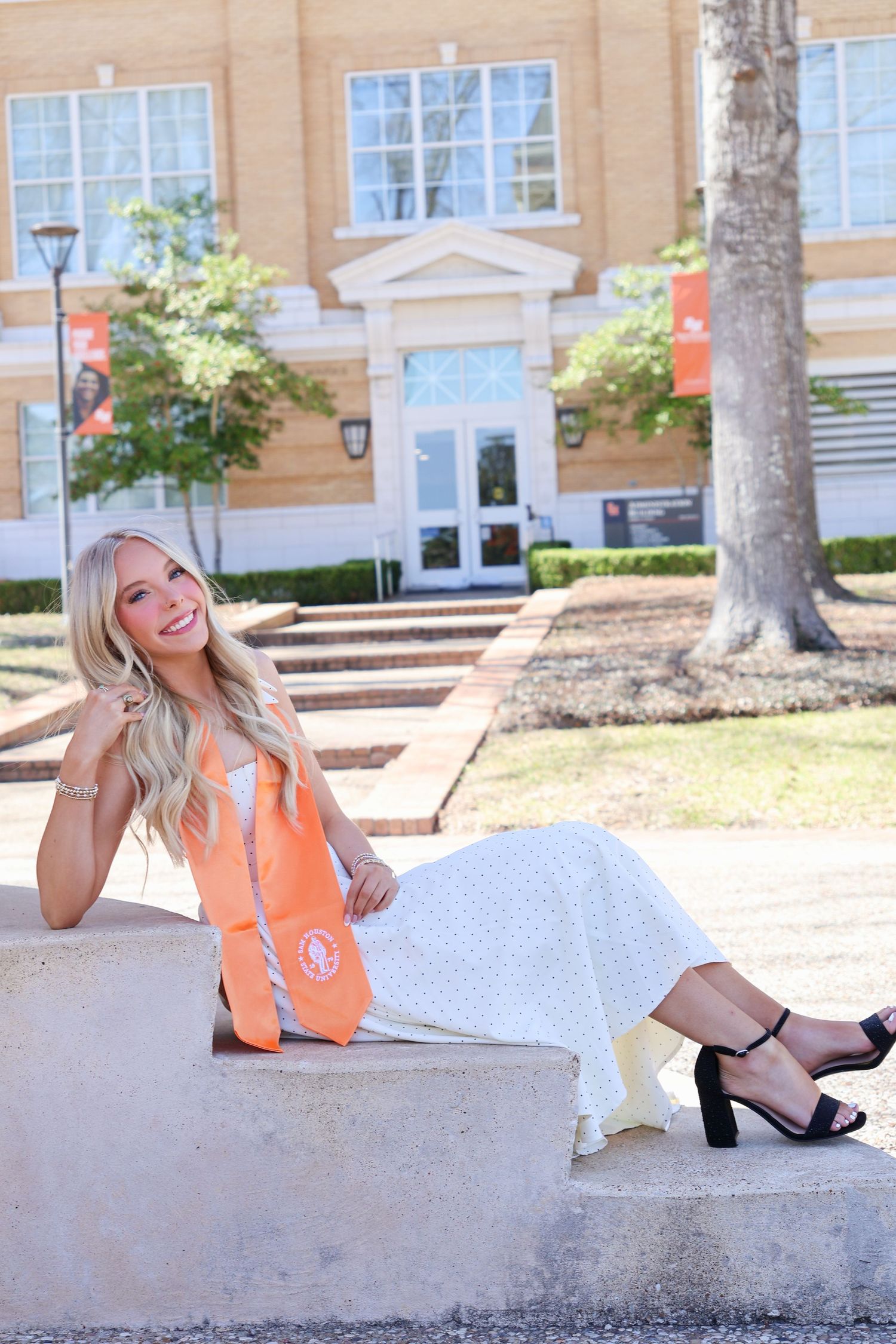 Smiling blonde graduate in white dress with orange graduation stole sits on stone wall outside university building.