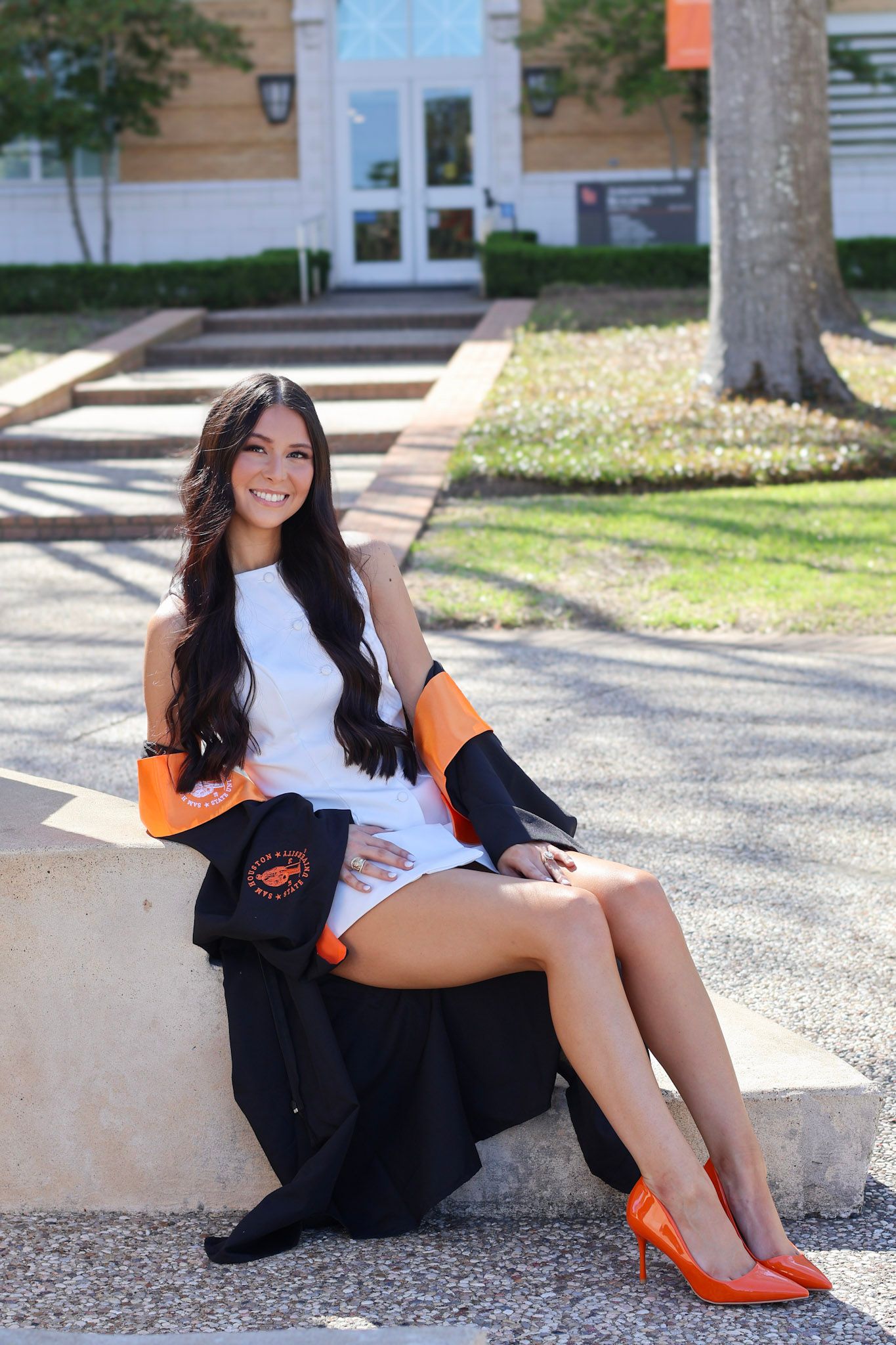 Young woman in white dress with orange graduation regalia sitting outdoors on campus steps smiling.