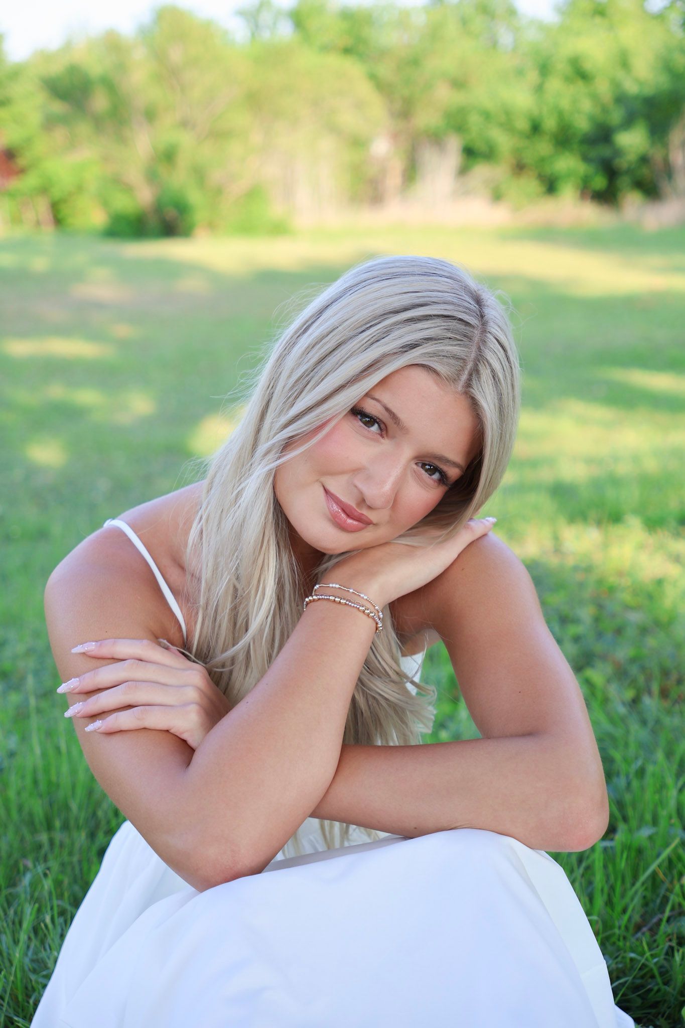 Blonde woman in white dress sitting on grass, resting head on hand, smiling softly.
