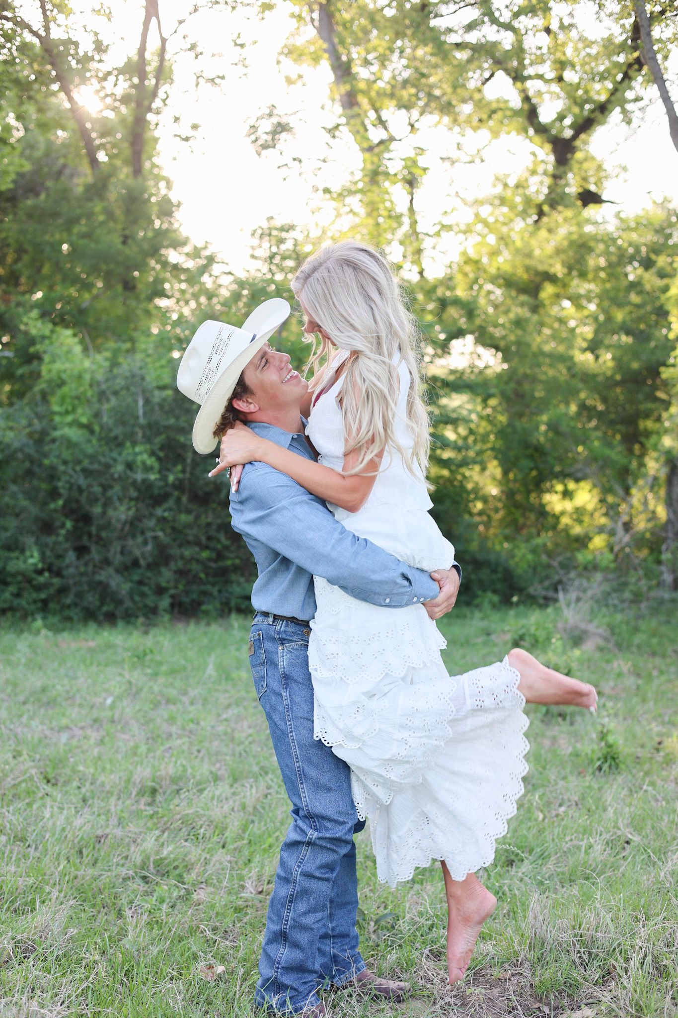 Couple sharing a romantic kiss in a green field, man in cowboy hat lifting woman in white lace dress.