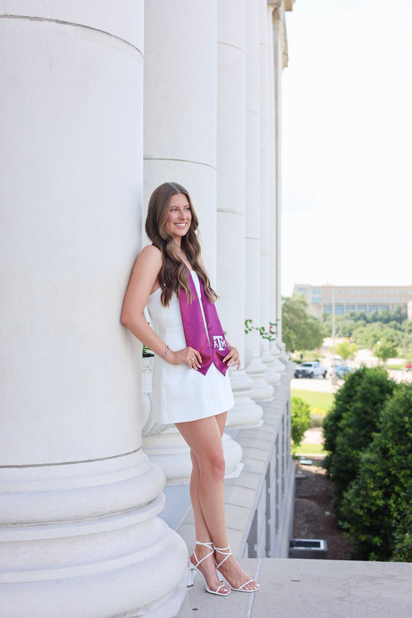 Young woman in white dress with Texas A&M maroon graduation stole leaning against white columns outdoors.