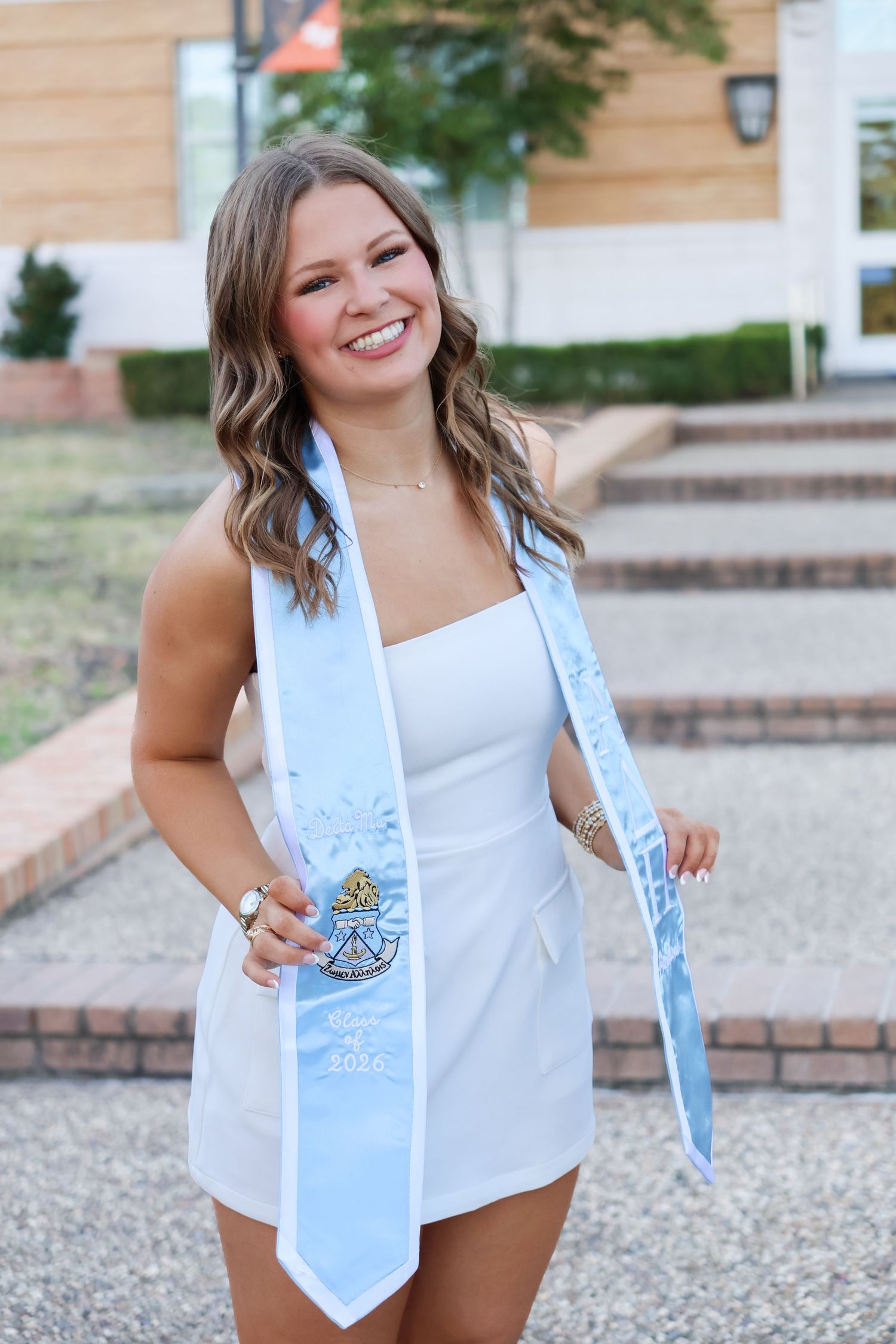 Smiling young woman in white dress wearing a light blue graduation stole, posing outdoors for graduation photos.