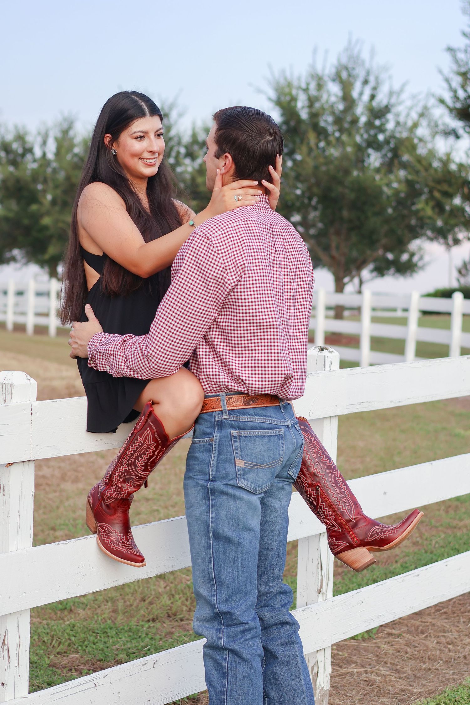 A couple shares a romantic moment by a white fence, with the woman wearing boots and a black top leaning against the railing.