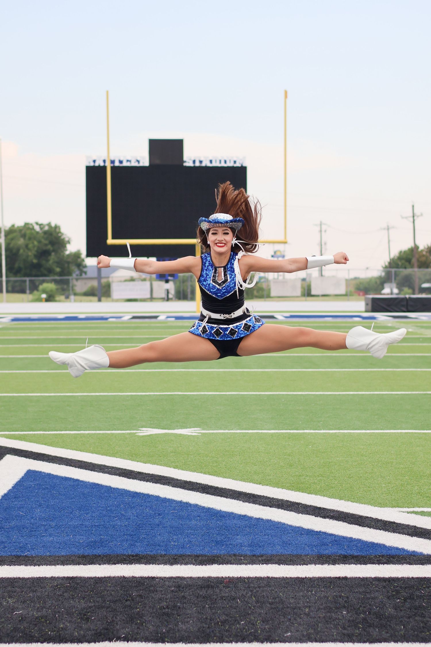 A cheerleader performs a split jump in mid-air on a football field with goal posts visible in the background.