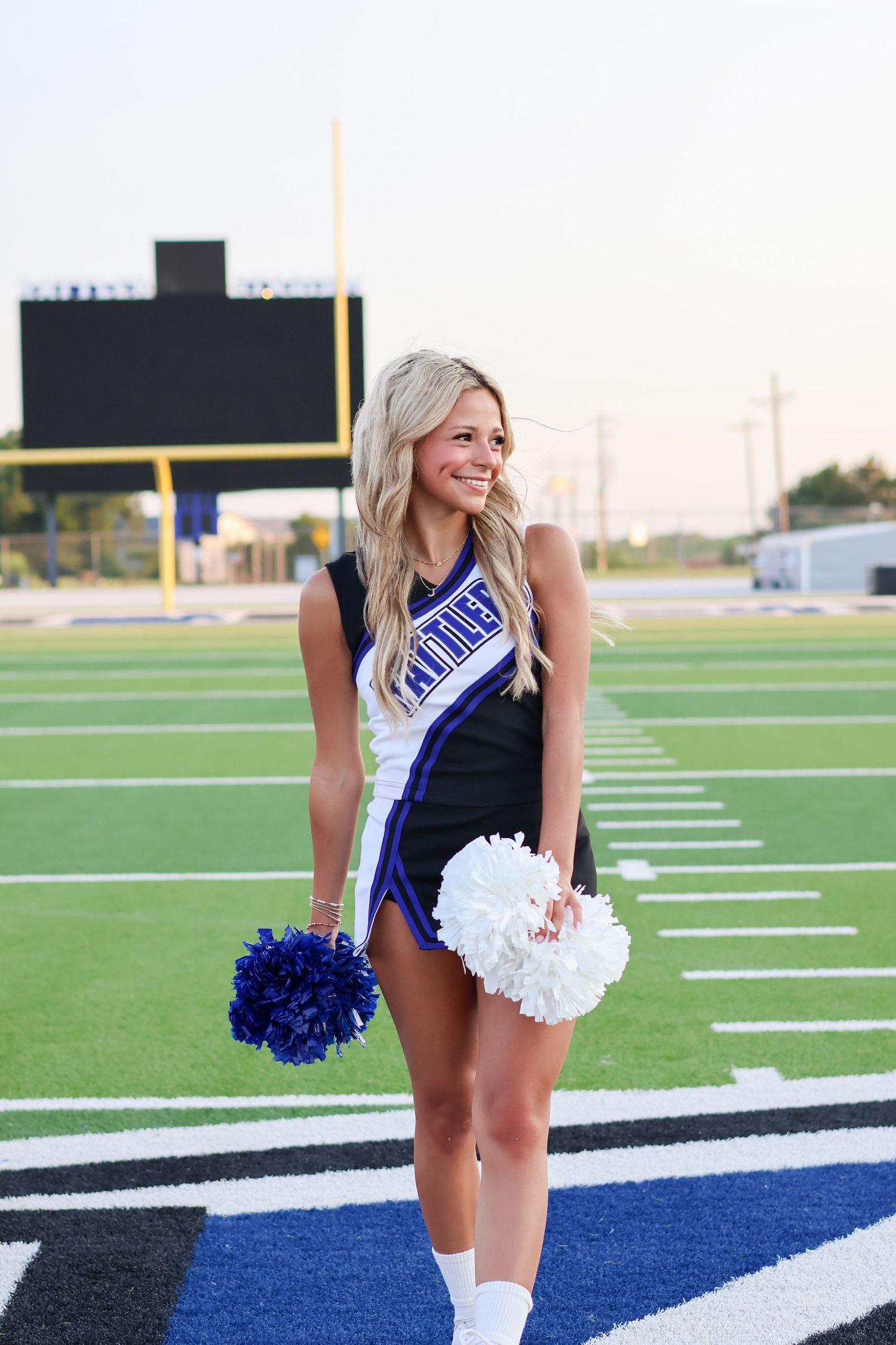 A cheerleader in blue and white uniform holding pom poms poses on a football field.