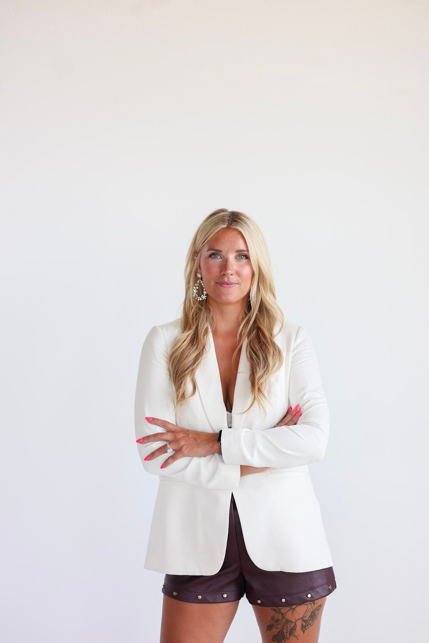 Confident blonde woman in white blazer and shorts with tattoo, arms crossed, posing against white background.