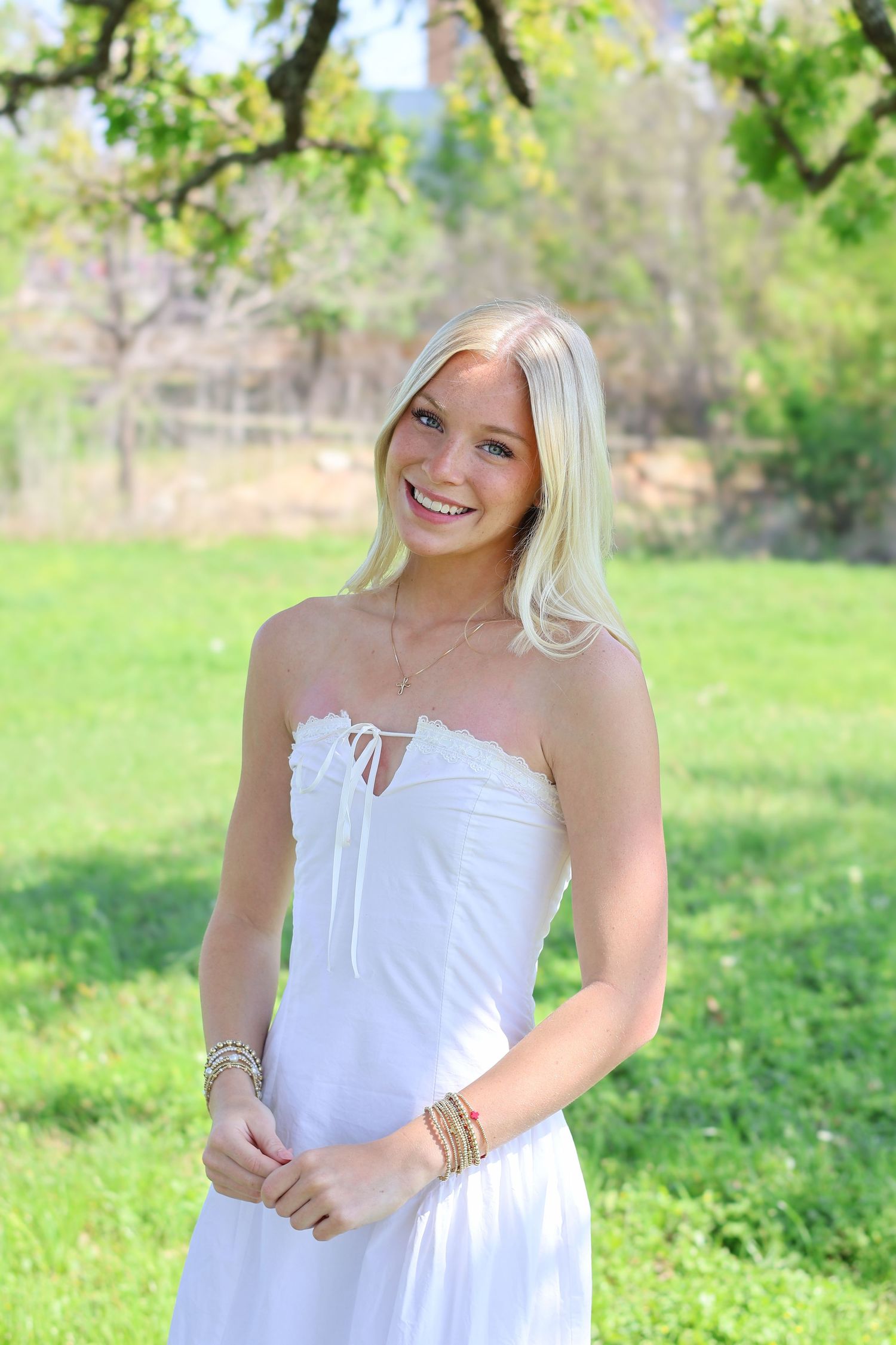 Smiling blonde woman in white strapless dress with bracelets standing outdoors in green park setting.