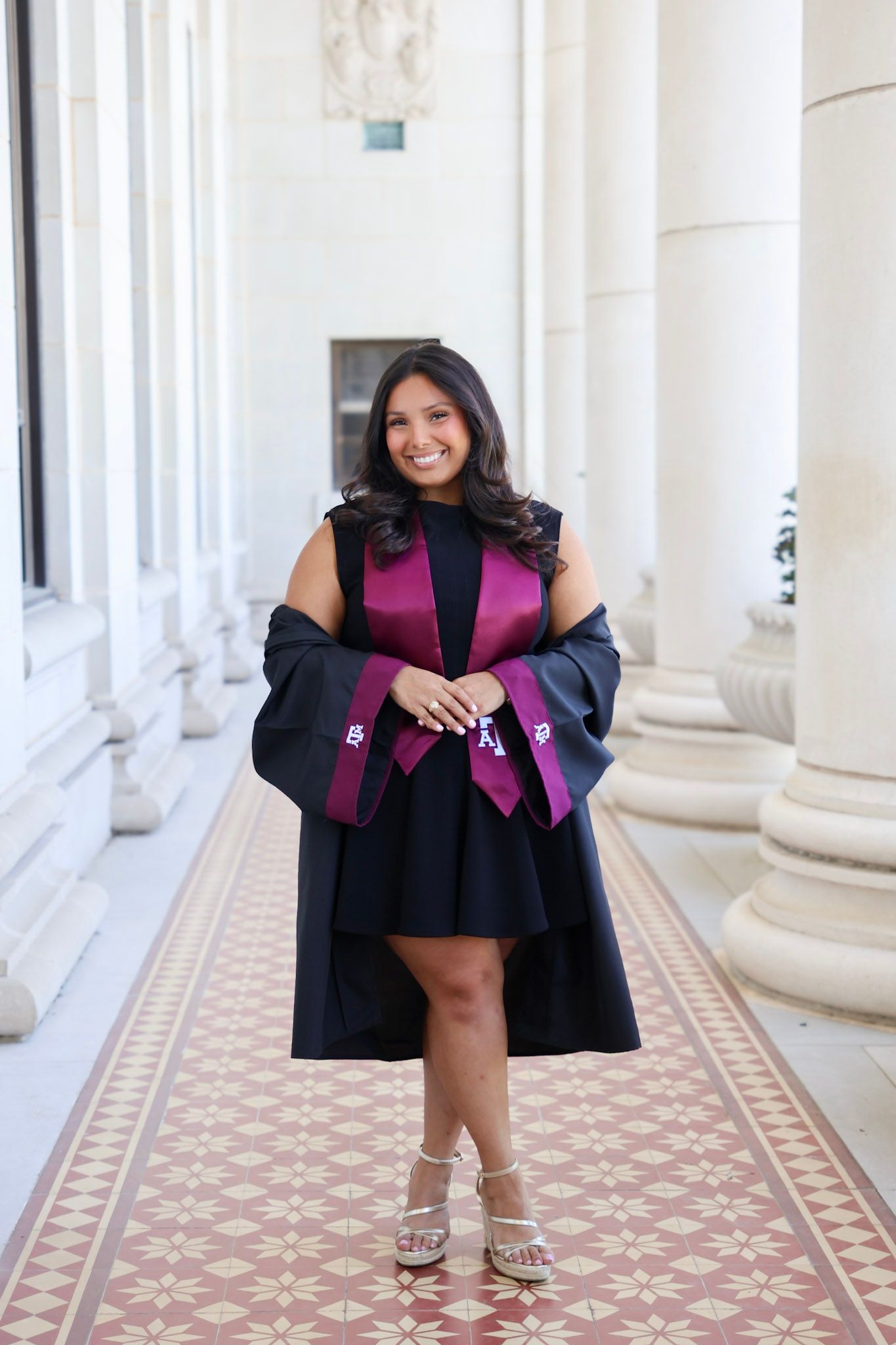 Smiling graduate in black gown with purple sash poses between white columns on decorative tiled floor.