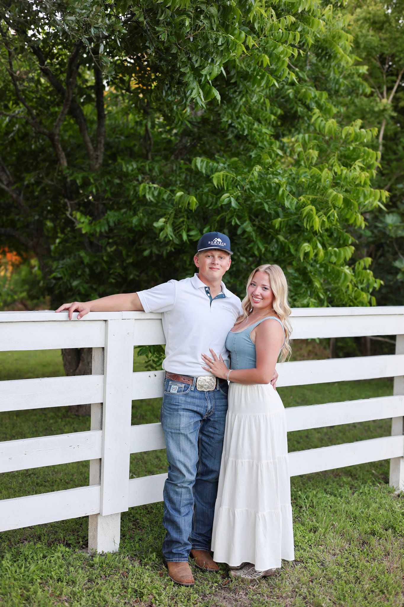 A couple poses by a white wooden fence surrounded by lush green trees, man in jeans and cap, woman in white skirt.